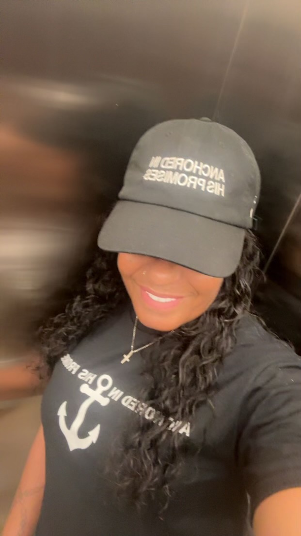 Person wearing a cap and black T-shirt with anchor design, smiling, in an elevator with dark walls.