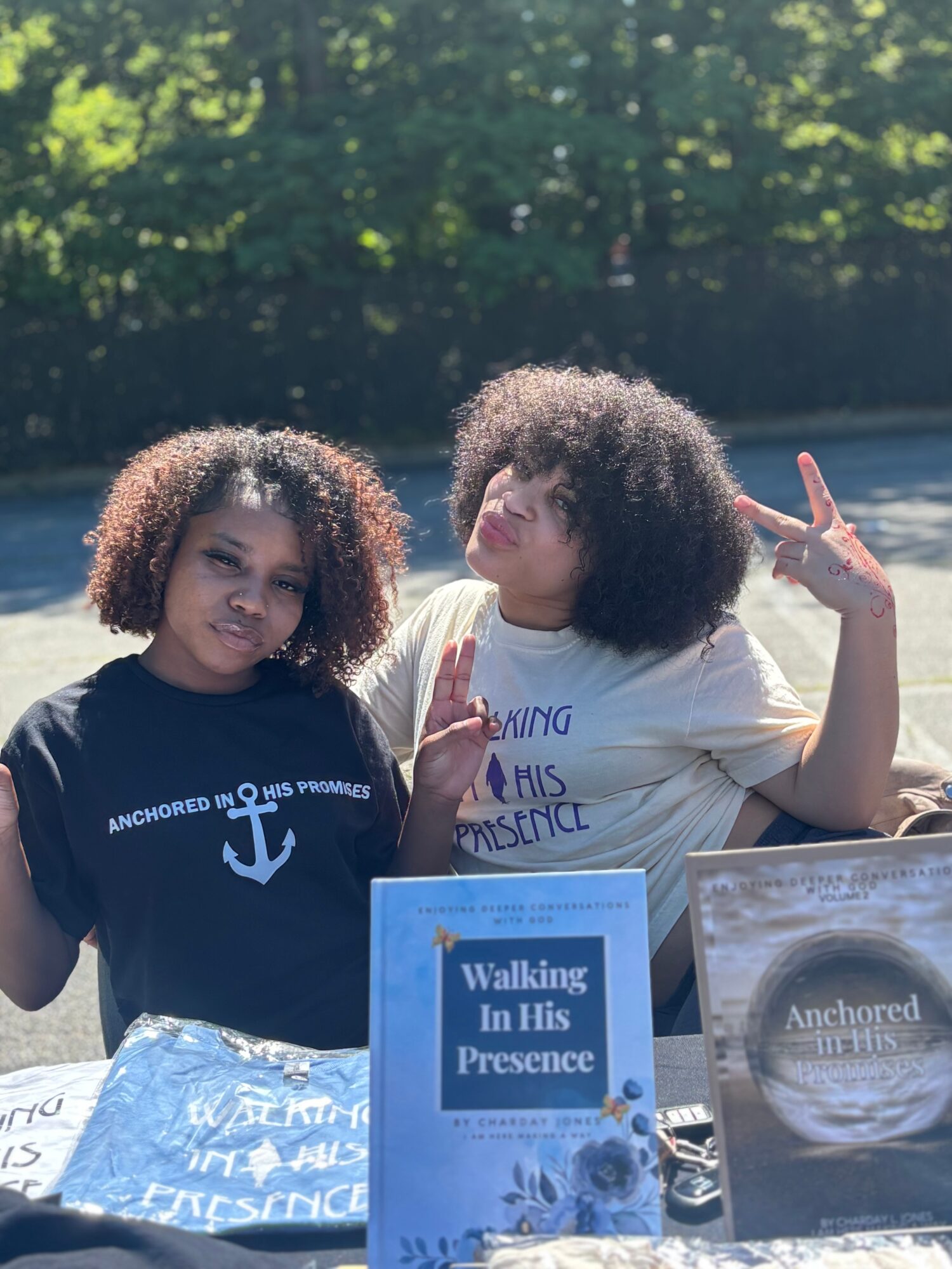 Two young women with curly hair standing outdoors behind a table with books, one making a peace sign, sunny day.