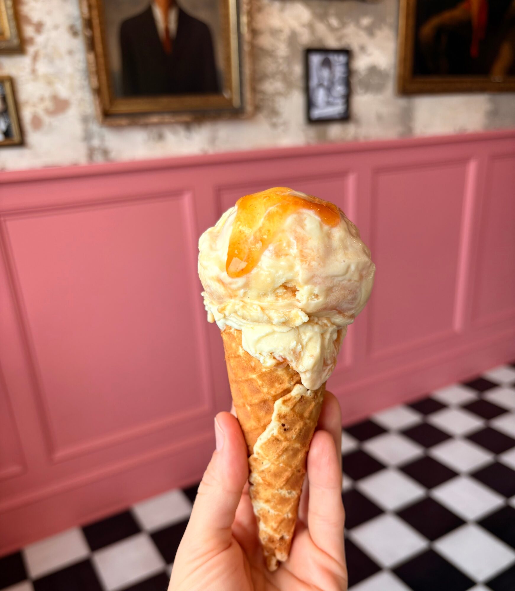 Hand holding an ice cream cone with two scoops, in front of a pink wall with framed pictures, black and white checkered floor.