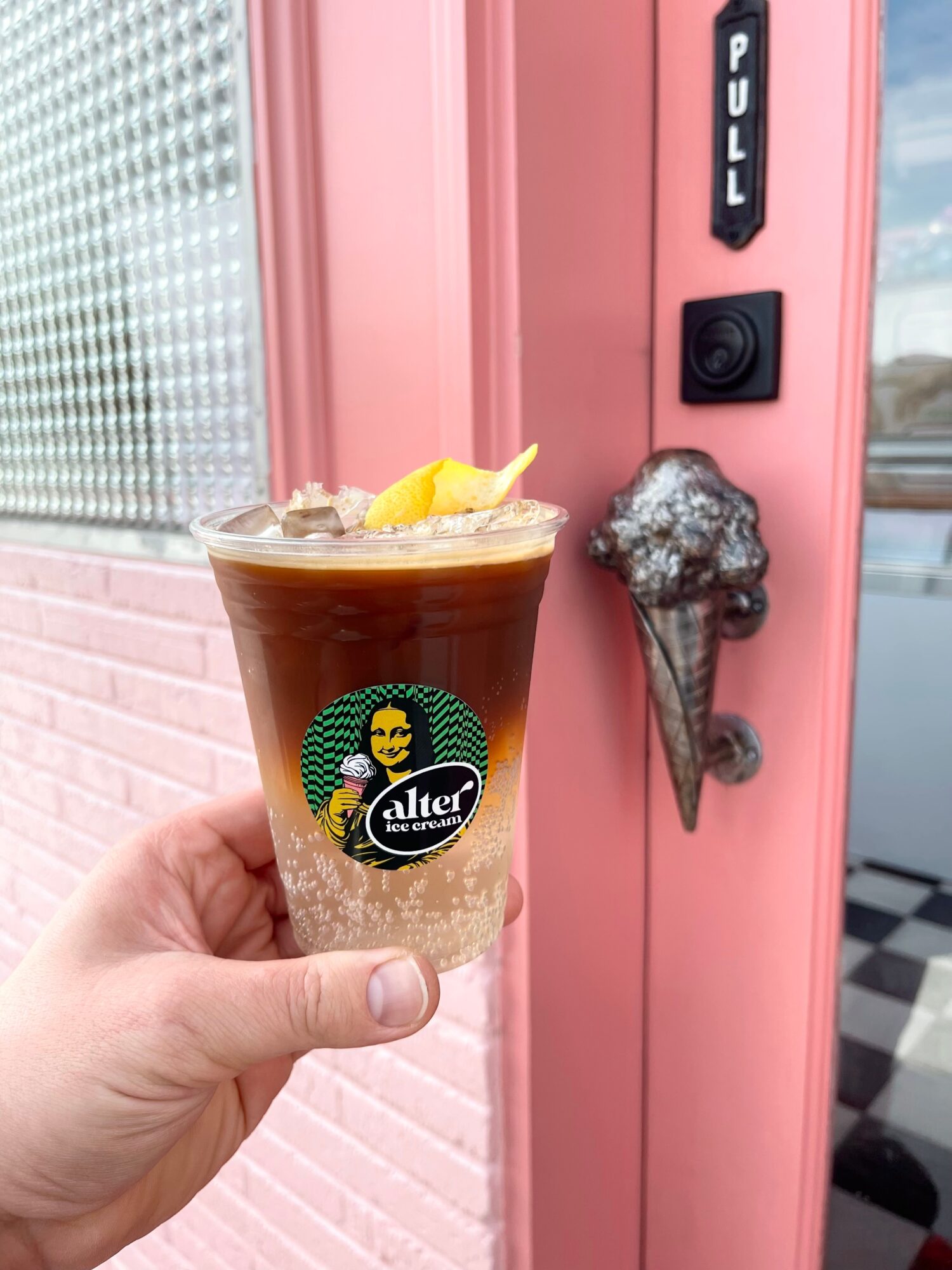 Hand holding a coffee drink with lemon peel, in front of a pink door with a black doorbell and ice cream cone decoration.