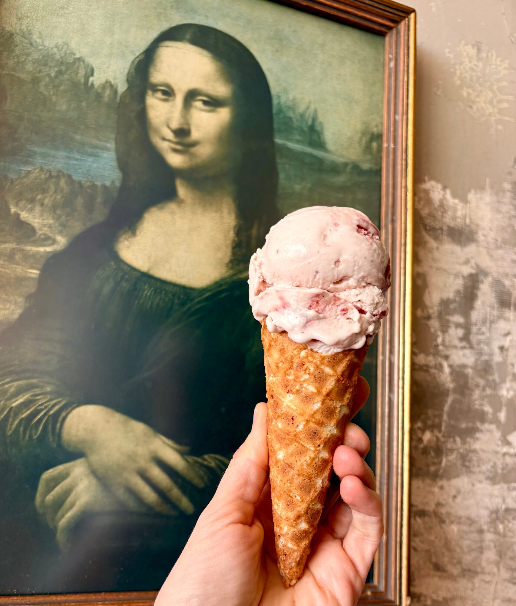 Person holding ice cream cone in front of Mona Lisa painting, with a cloudy sky background, indoors.