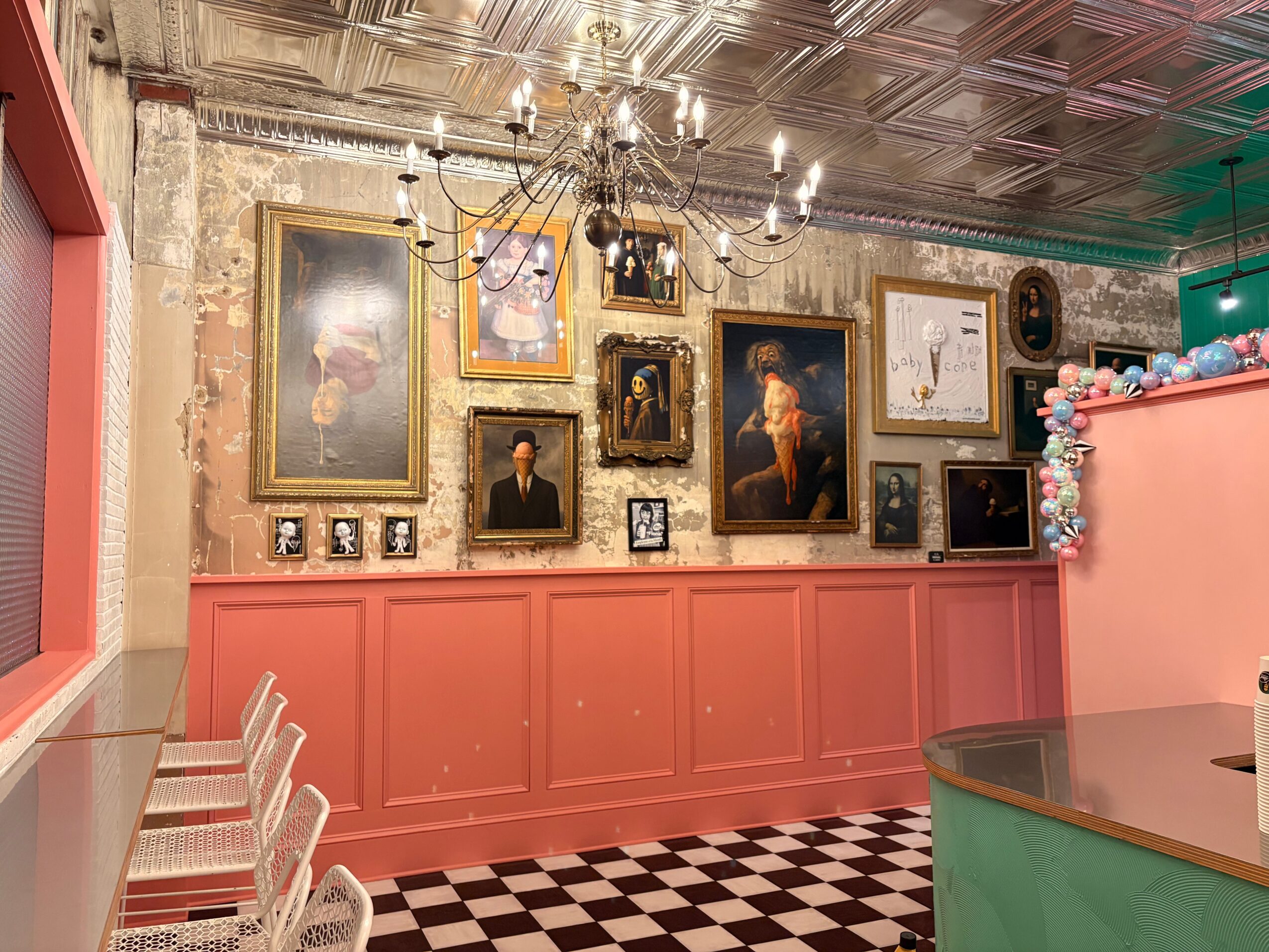 Interior with a pink wall, framed paintings, a chandelier, and checkered floor tiles, with a counter on the right side.