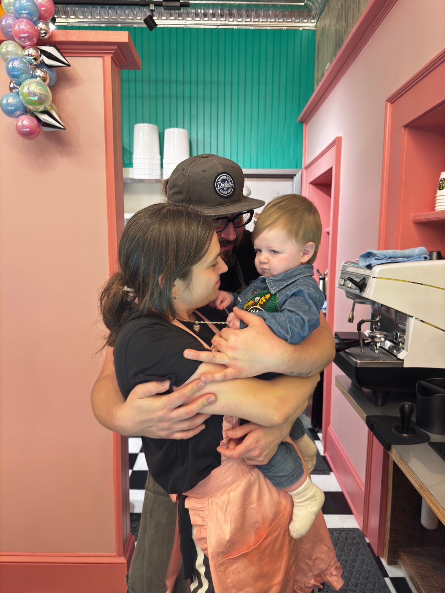 Child and woman hugging in a colorful room with pink walls and balloons, woman wearing a black shirt and child in denim.