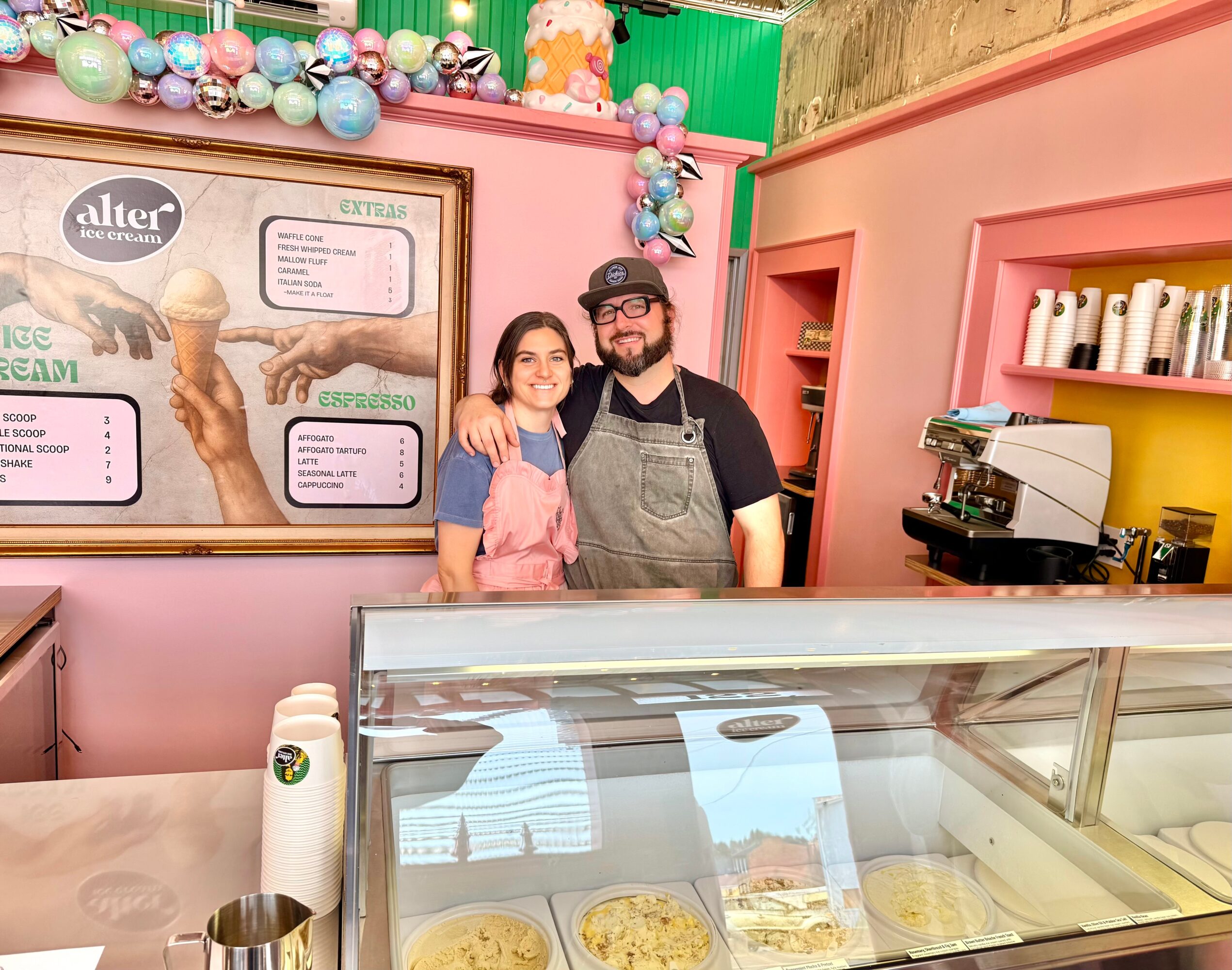 Two people stand behind an ice cream counter, smiling, with colorful decorations and a pink wall in the background.