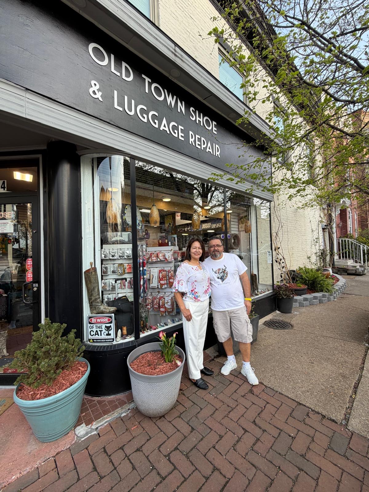 Two people stand outside a shoe and luggage repair shop on a sidewalk, smiling at the camera.