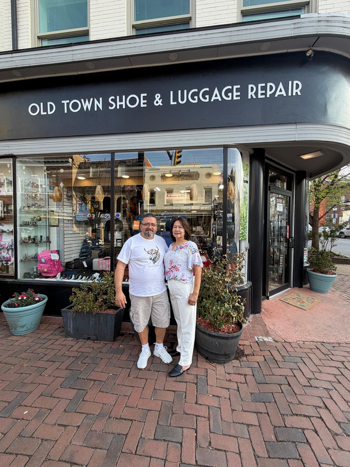 Two people stand outside a shoe and luggage repair shop with large windows and potted plants, smiling at the camera.
