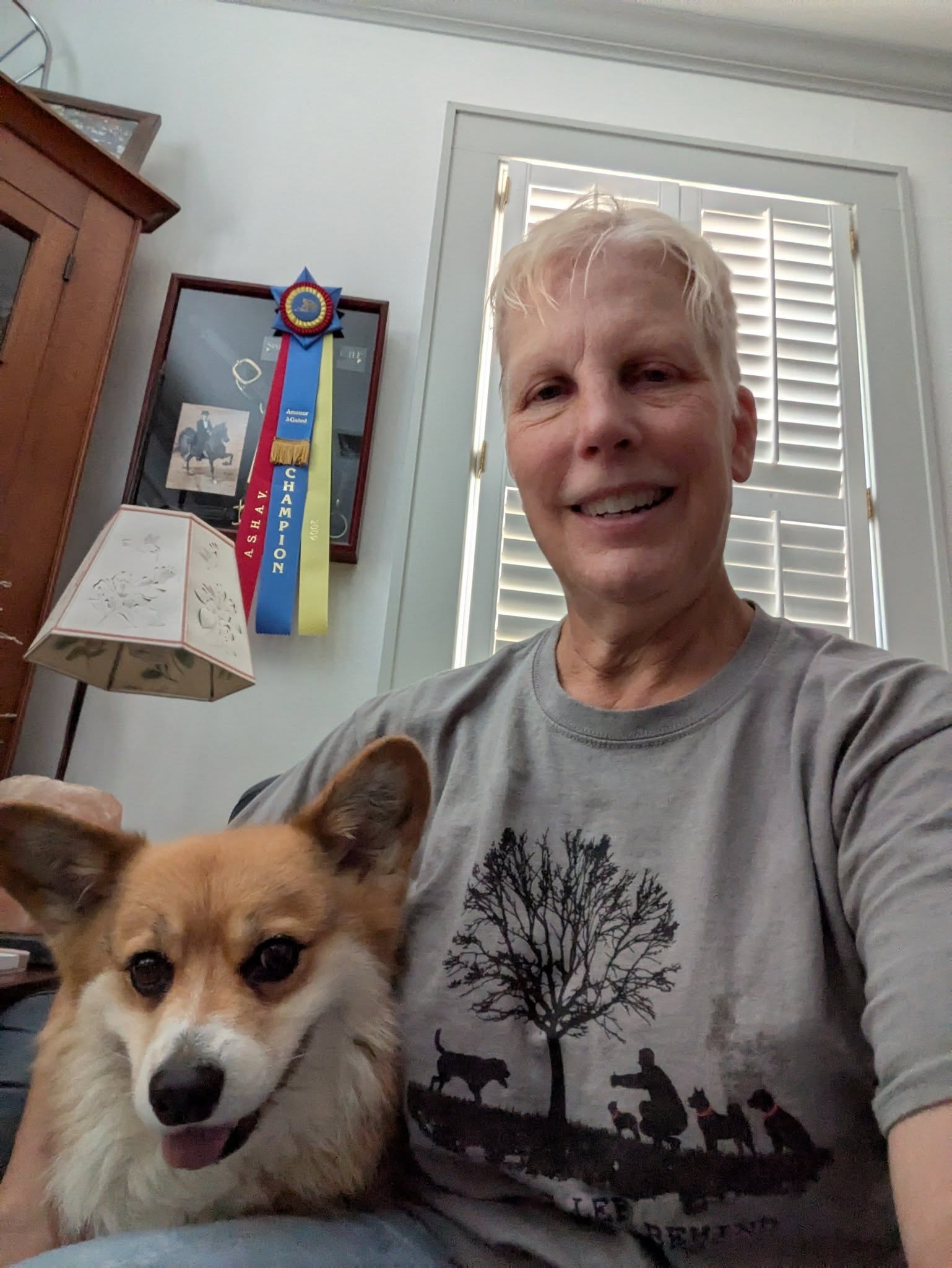 Smiling woman with short blonde hair taking a selfie with a small dog indoors, window with shutters in background.