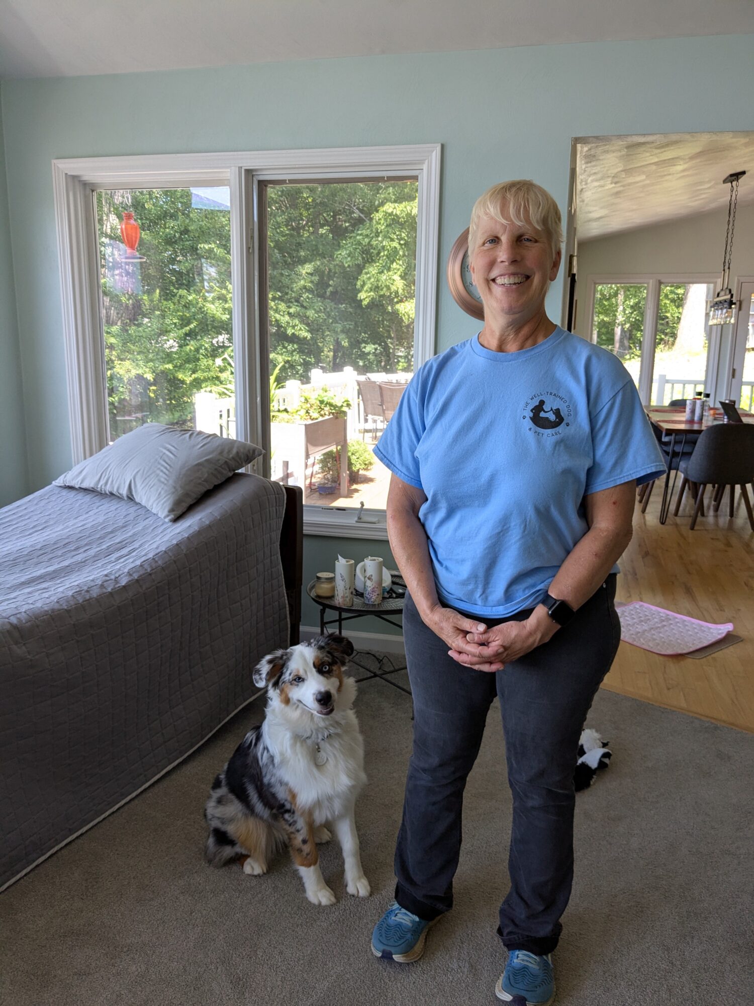 Woman with short blonde hair smiling, standing indoors near a dog, in a room with large windows and a view outside.