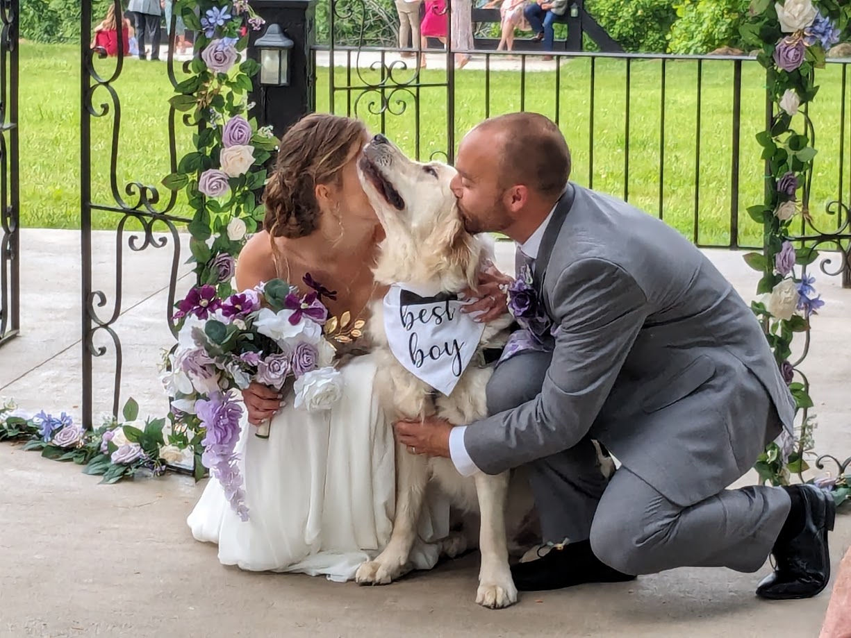 A couple and a dog at a wedding ceremony outdoors, with floral decorations and a black metal gate in the background.