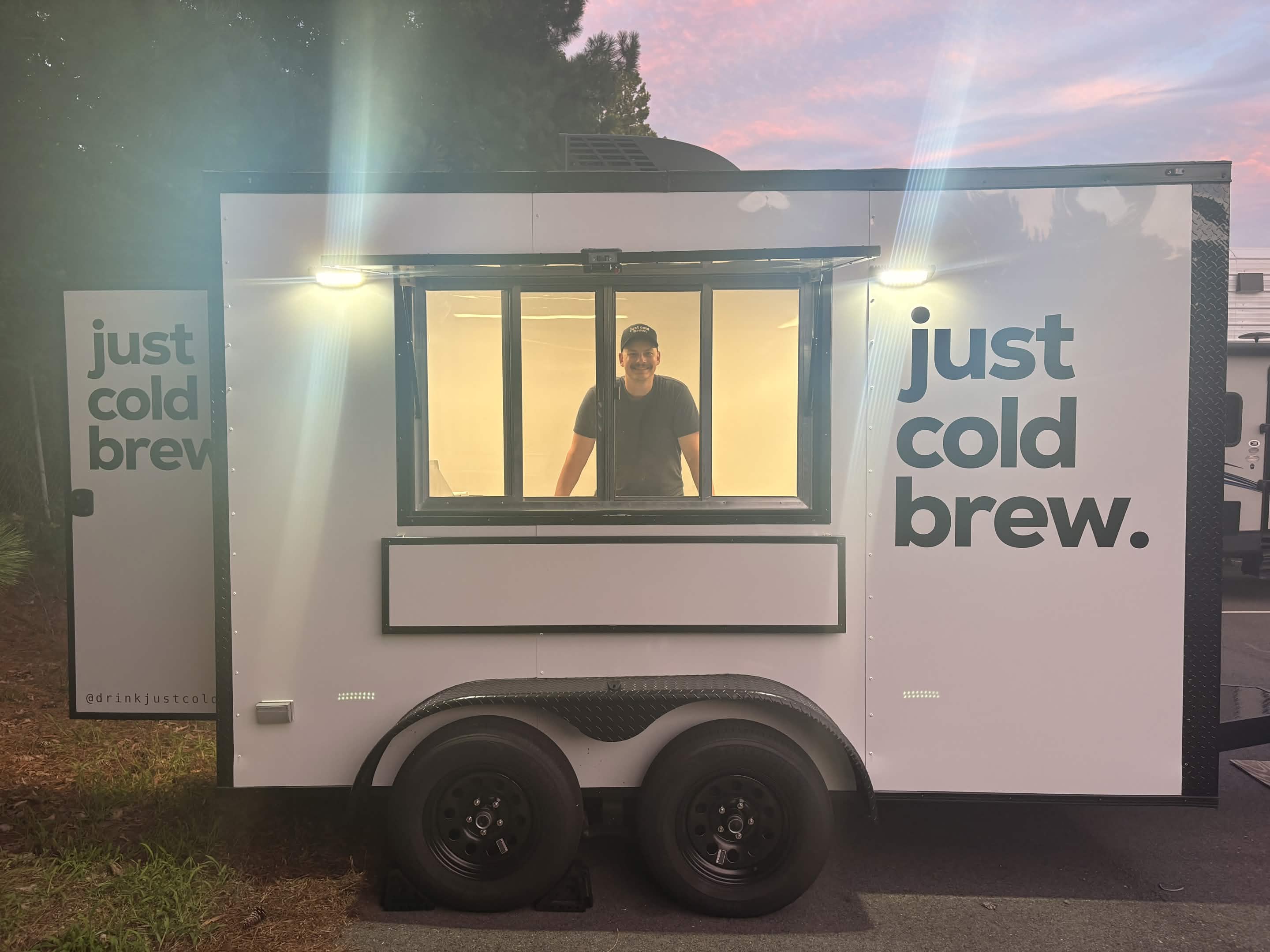 Person standing inside a small mobile coffee stand with 'just cold brew' signs, two tires below, and a window with a person inside.
