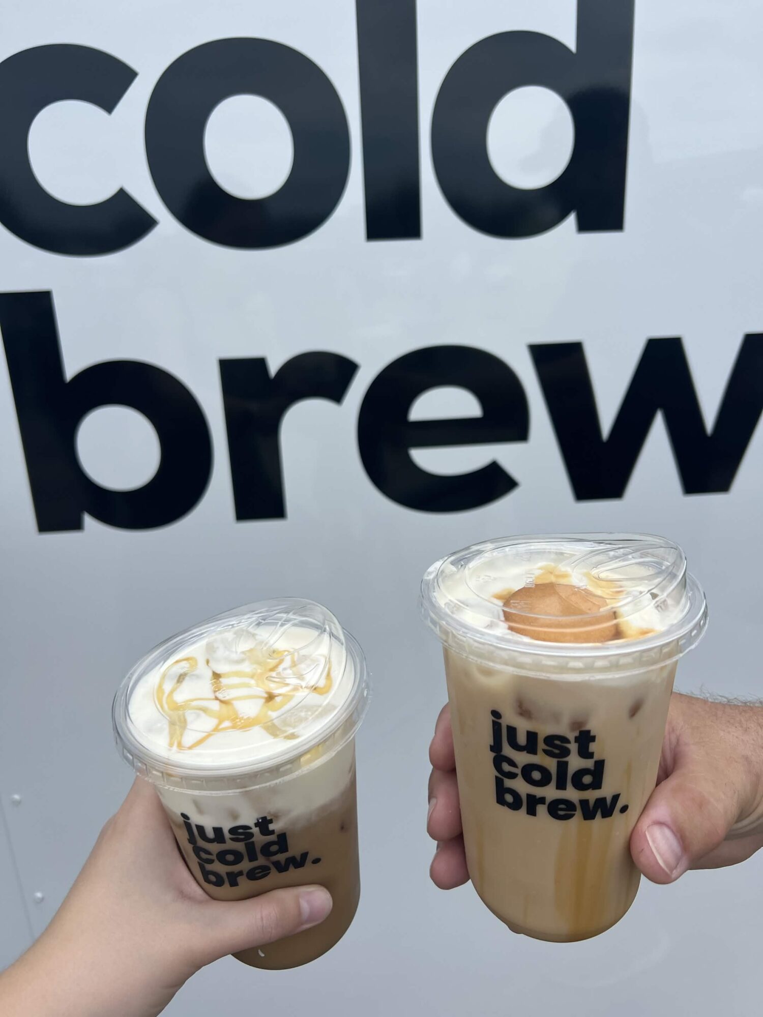 Two cups of cold brew coffee held up in front of a sign that says 'cold brew'.