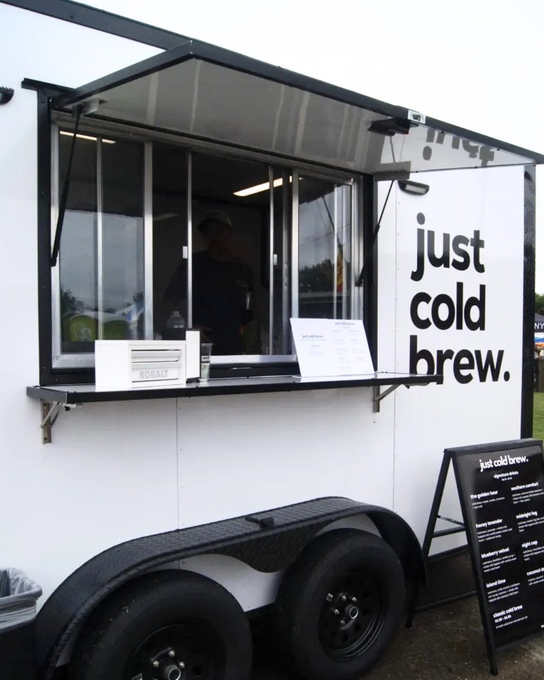 A mobile coffee stand with a sign that says 'just cold brew,' featuring a menu board outside.