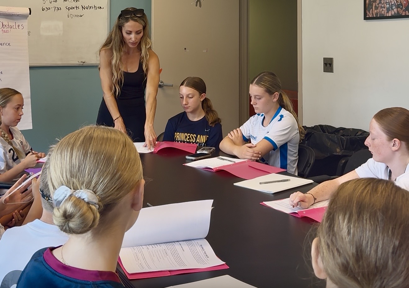 Group of young girls and a woman in a classroom, sitting around a table with papers and folders, engaging in discussion.