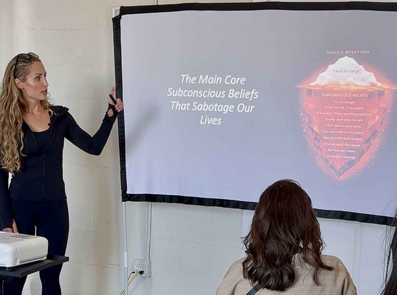 Woman presenting a slide with text about subconscious beliefs and lives, with a person in the audience, in a classroom or conference room.