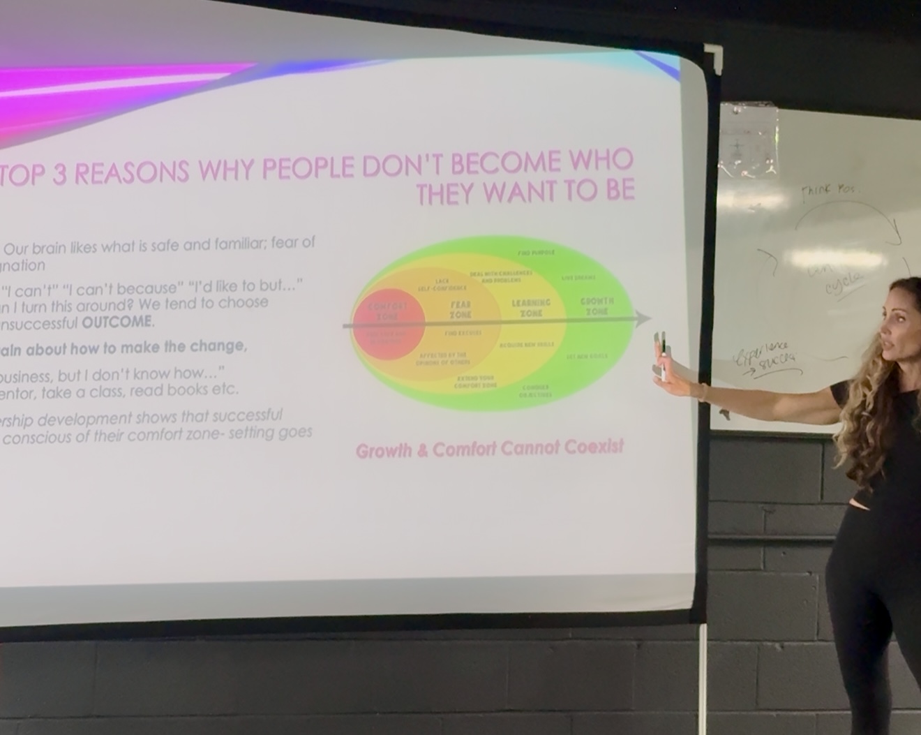 A woman presents a slide titled 'Top 3 reasons why people don't become who they want to be' with a colorful diagram.