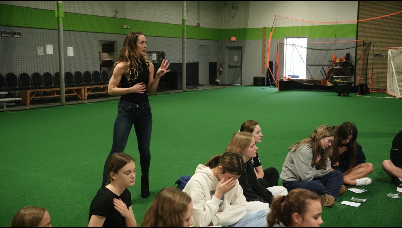 Group of young people sitting on the floor in a gym, with a woman standing and speaking, green floor and walls.