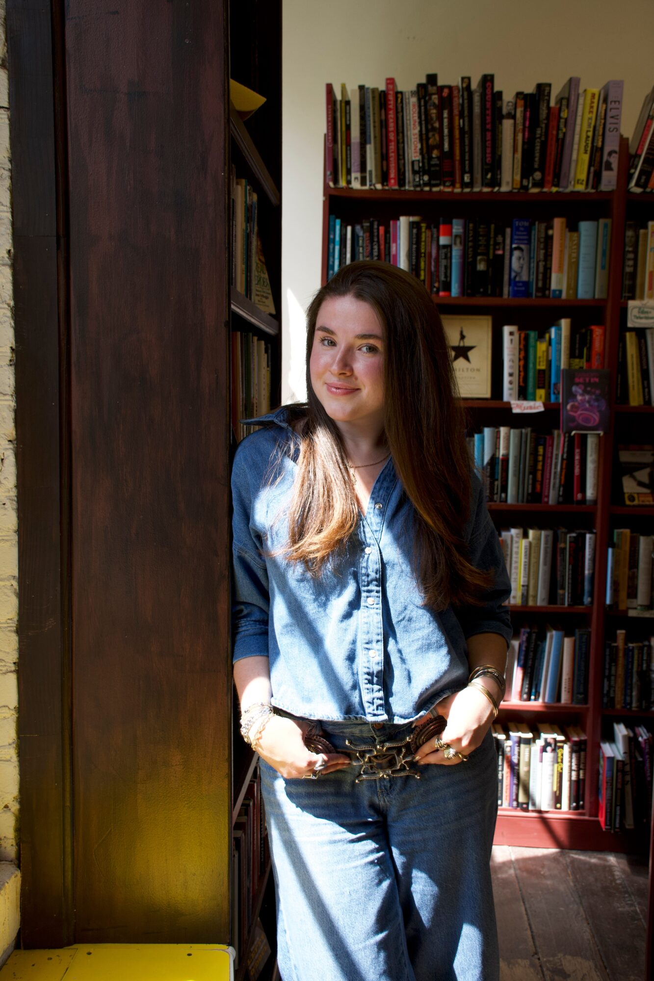 Young woman with long hair wearing a denim shirt and jeans, standing in front of a bookshelf in a library.