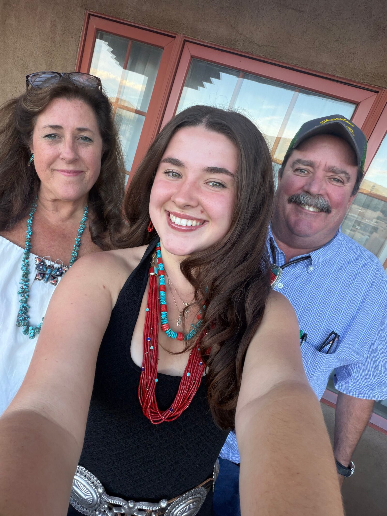Three people smiling outdoors, woman in center taking selfie, two others beside her, background with windows and building.