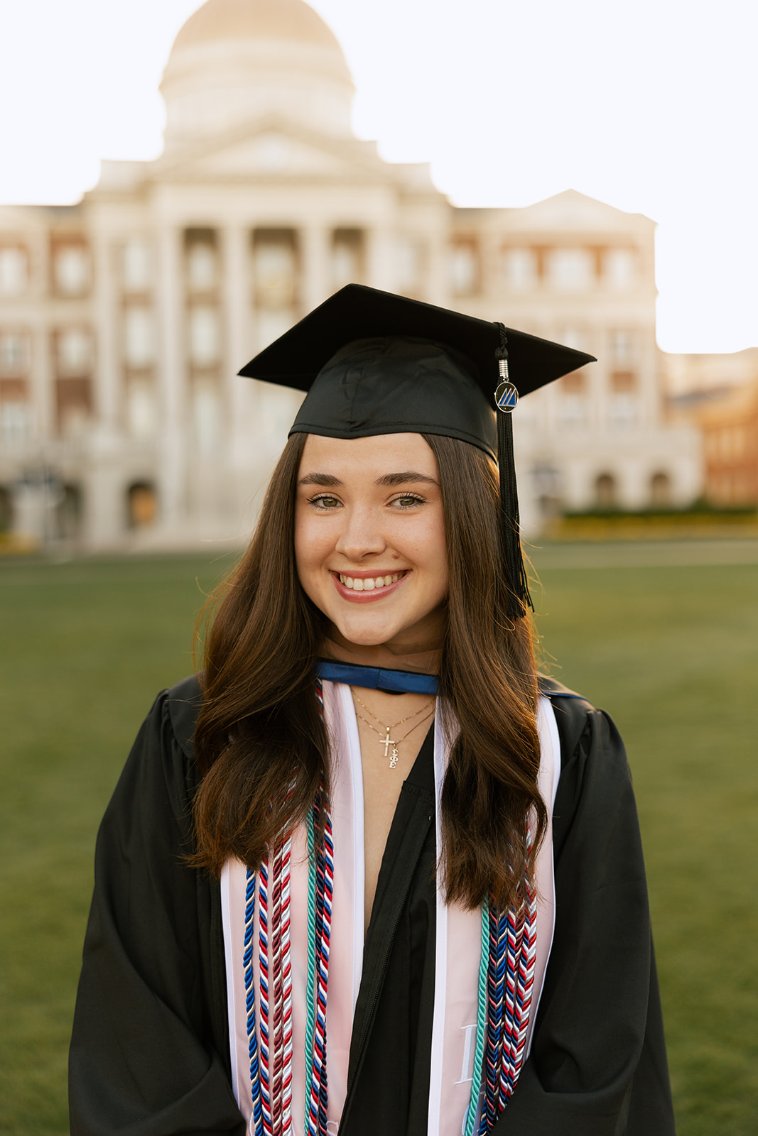 Young woman in graduation gown and cap smiling outdoors in front of a large building.