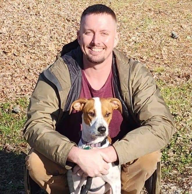 Man smiling outdoors holding a small dog, both facing camera, sunlight on face and ground, background of dry grass and leaves.
