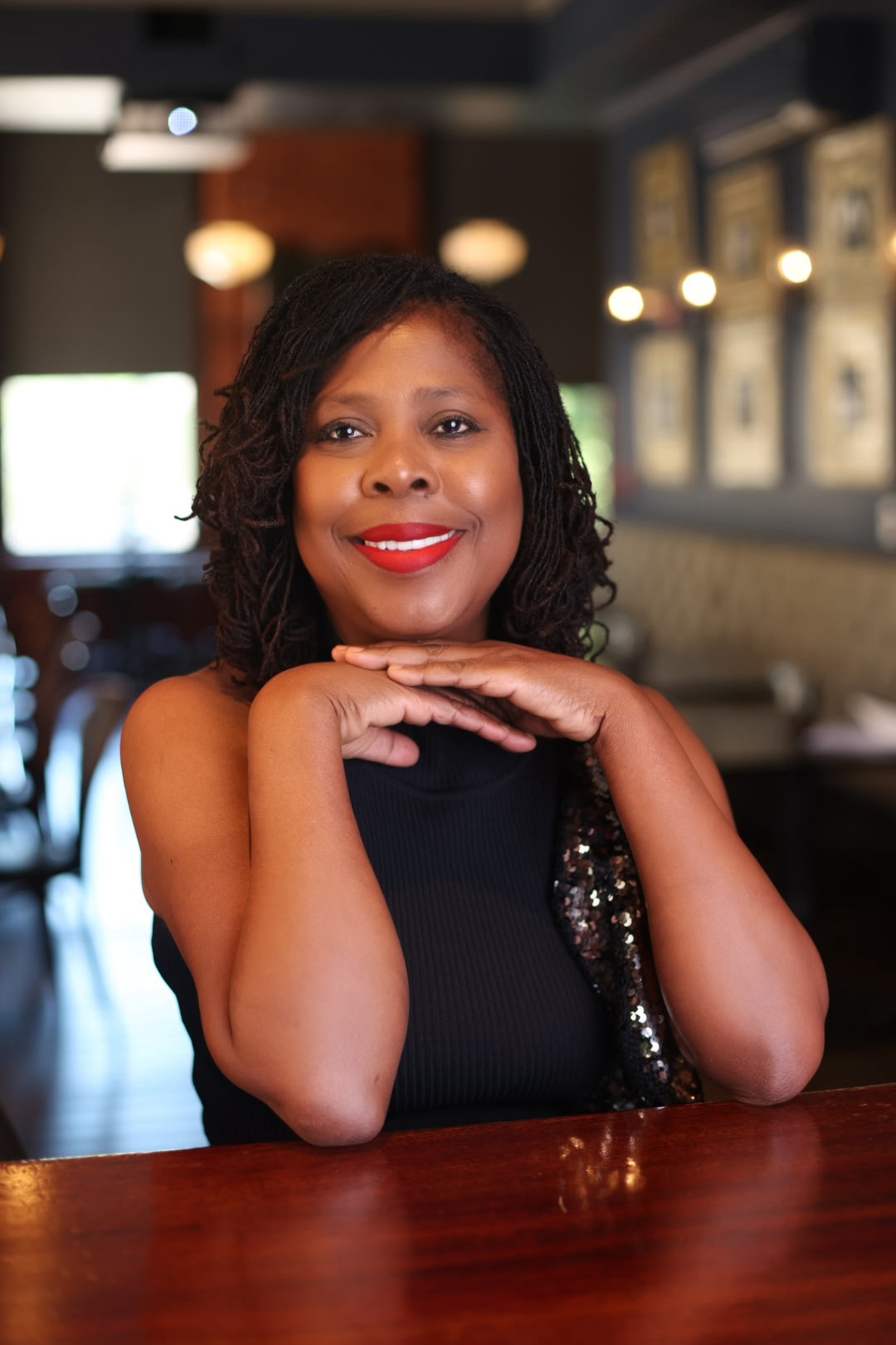 Woman with curly hair and red lipstick resting her chin on her hands, smiling at the camera in a cafe.