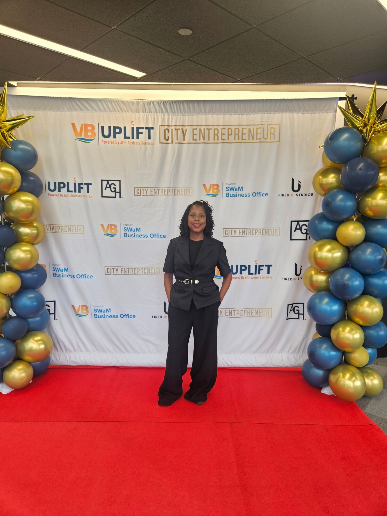 Woman in black suit standing on red carpet in front of a backdrop with balloons and event logos.