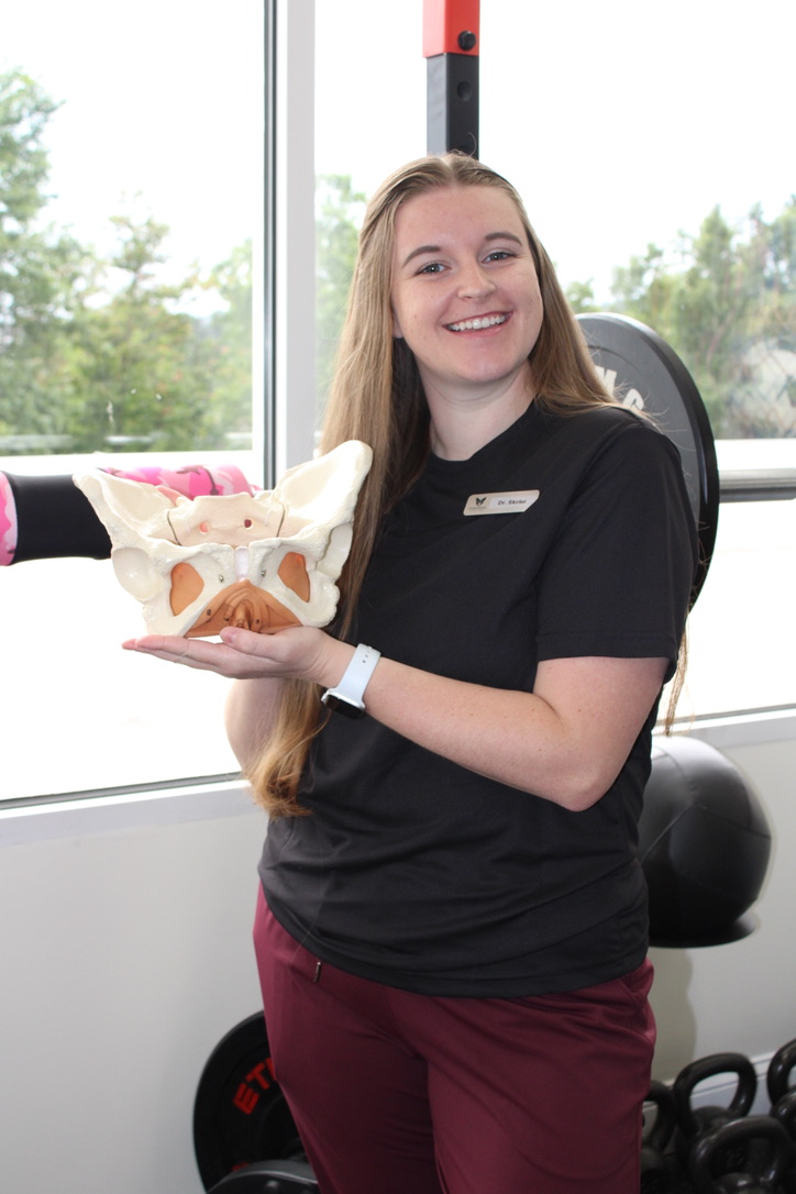 Young woman with long hair smiling, holding a model of a pelvis, in a fitness or therapy setting.