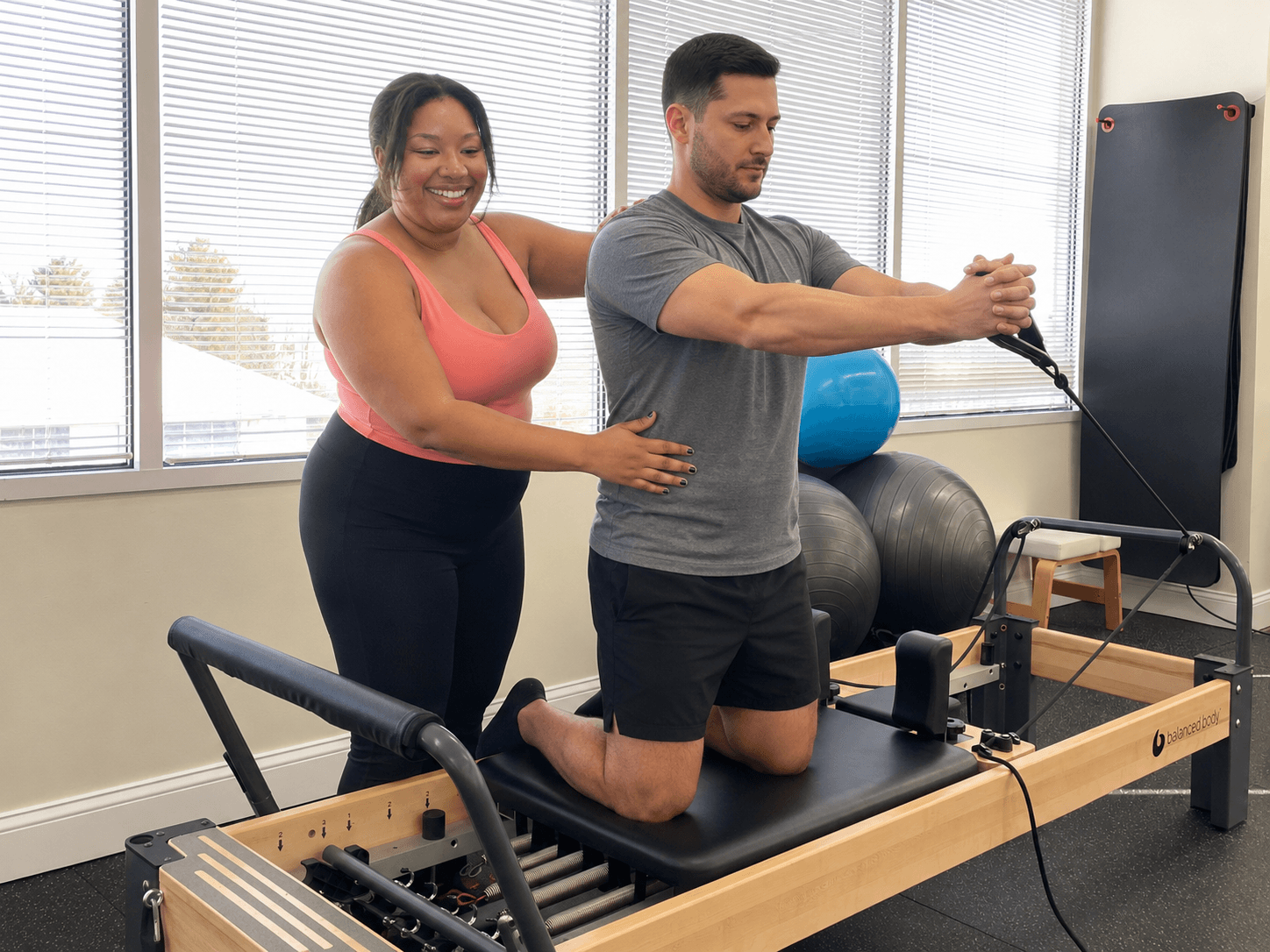A woman and a man are exercising on a reformer machine in a gym, with the woman smiling and the man focused.