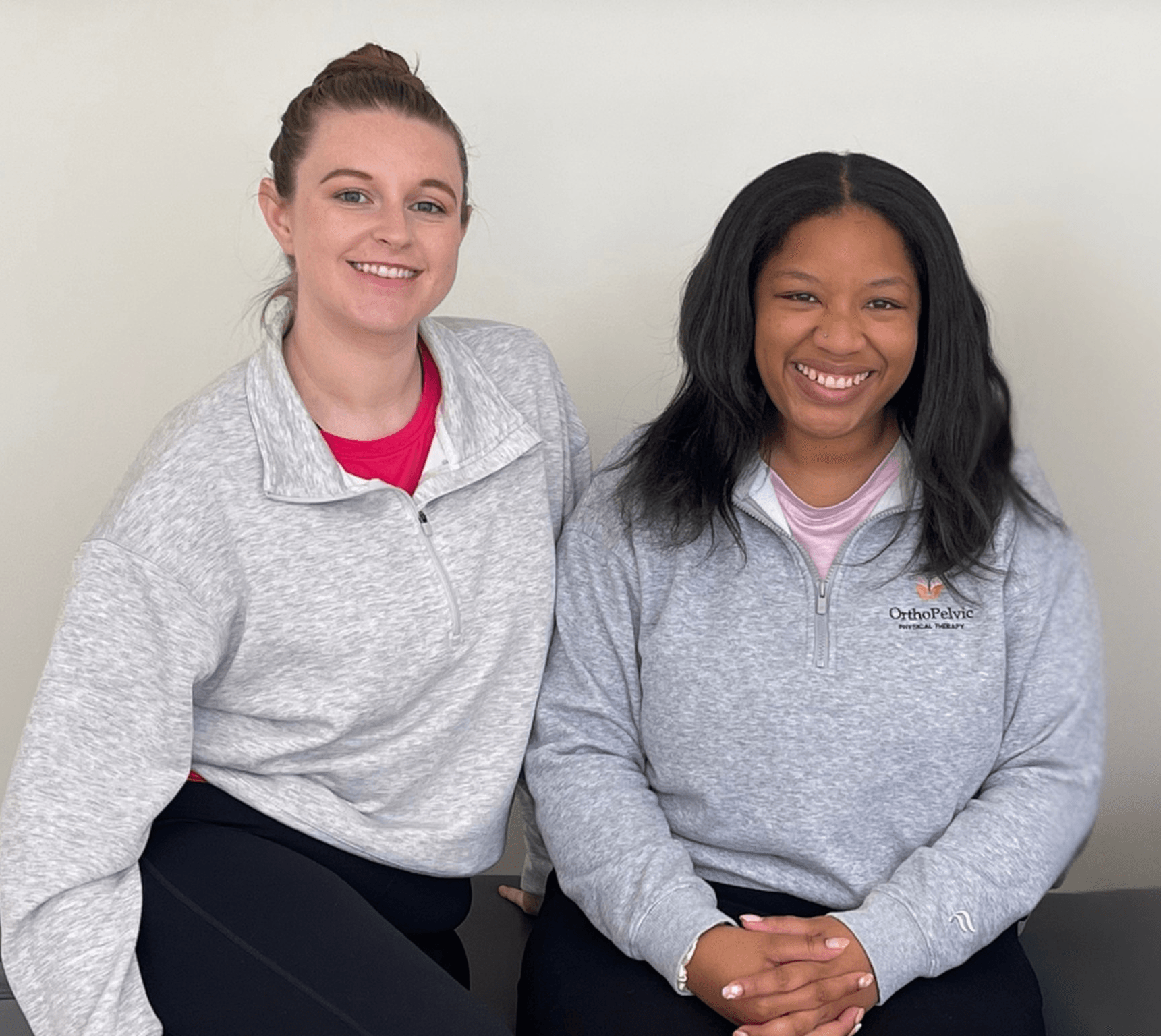 Two women smiling, standing side by side, wearing gray sweatshirts, against a plain white wall.