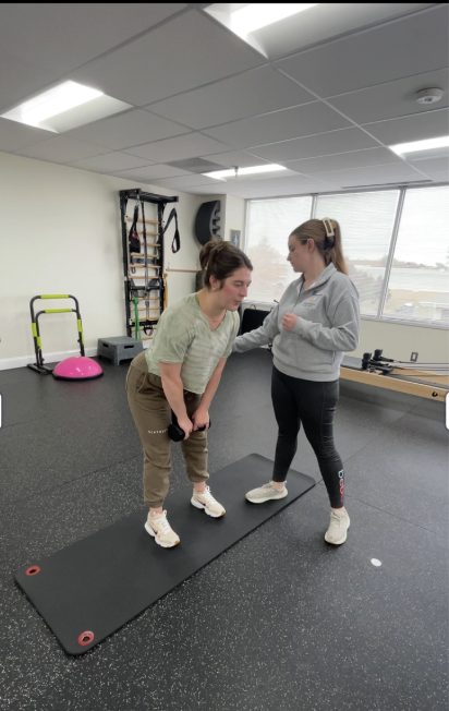 Two women stand on a black mat in a room with fitness equipment, engaging in a conversation or activity.
