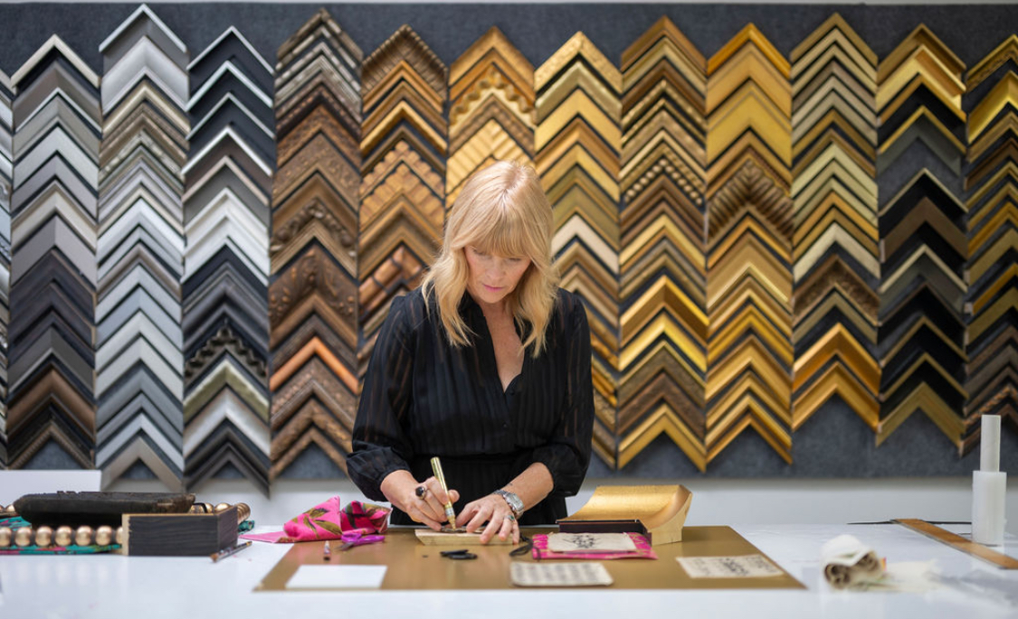 Woman with blonde hair in a black shirt writes on a piece of paper at a table in front of a wall of colorful picture frames.