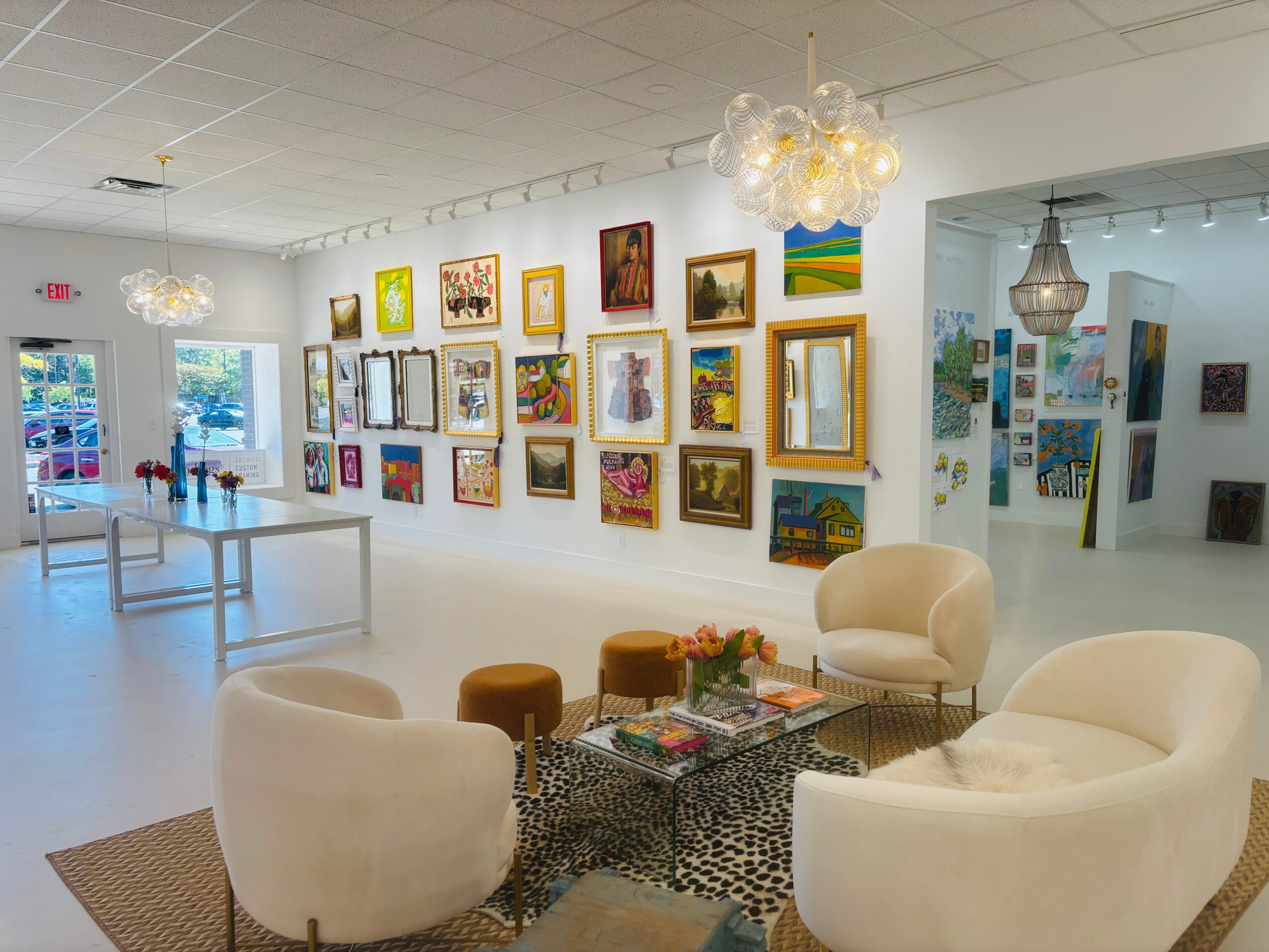 Art gallery with colorful paintings on white walls, seating area with white chairs, tables, and decorative lighting fixtures.