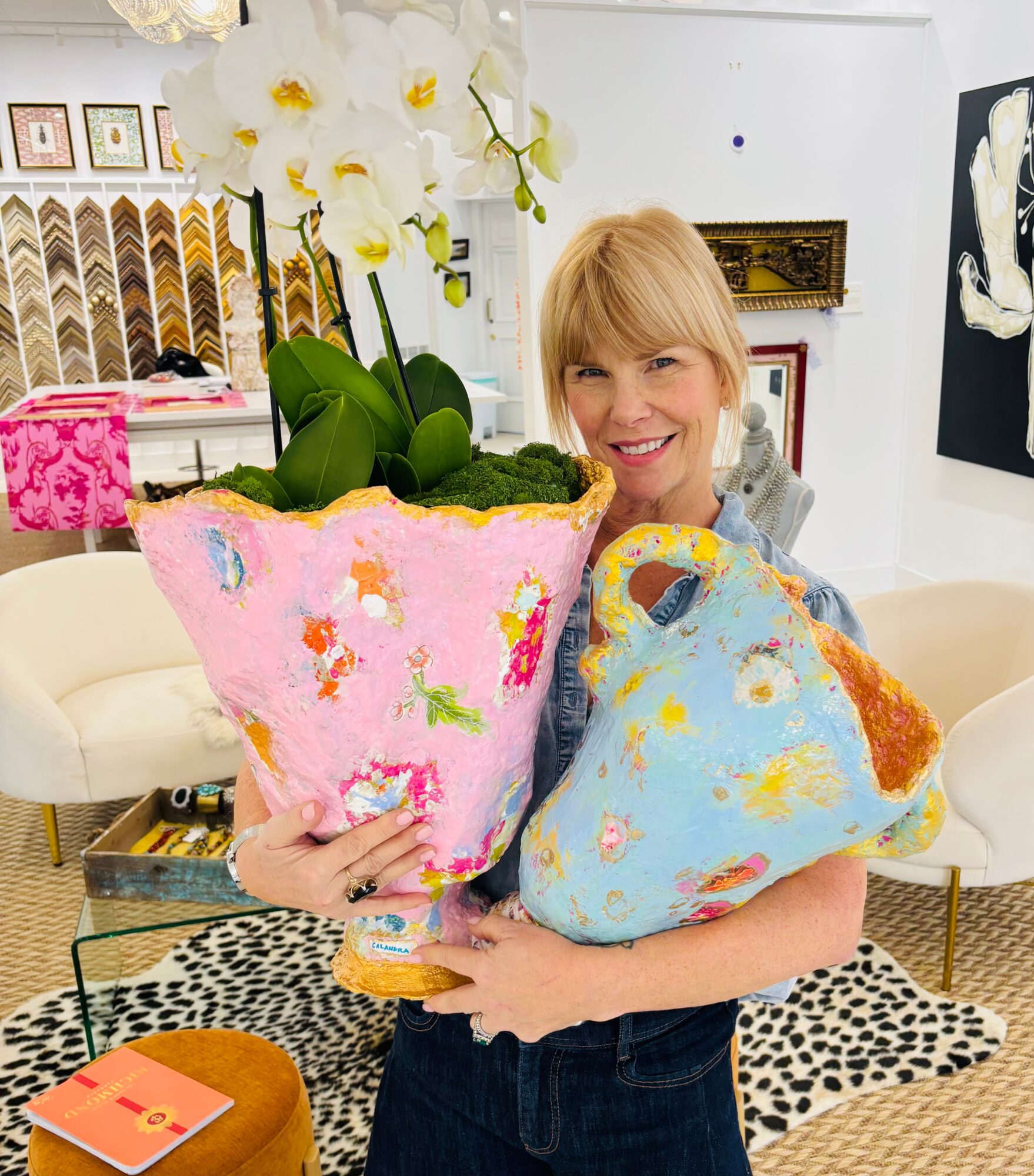 Woman smiling holding two colorful plush bags in a room with white walls, artwork, and a patterned rug.