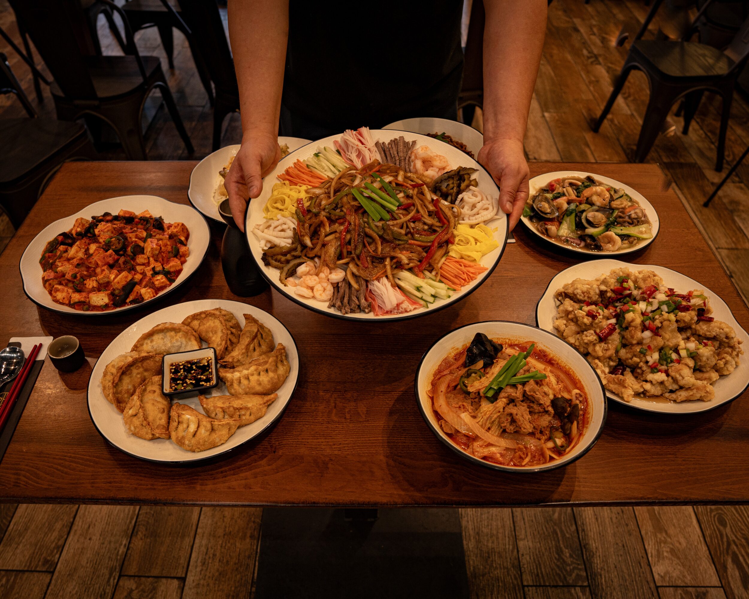 Person holding a large bowl of noodles with vegetables and meat, surrounded by six other dishes on a wooden table.