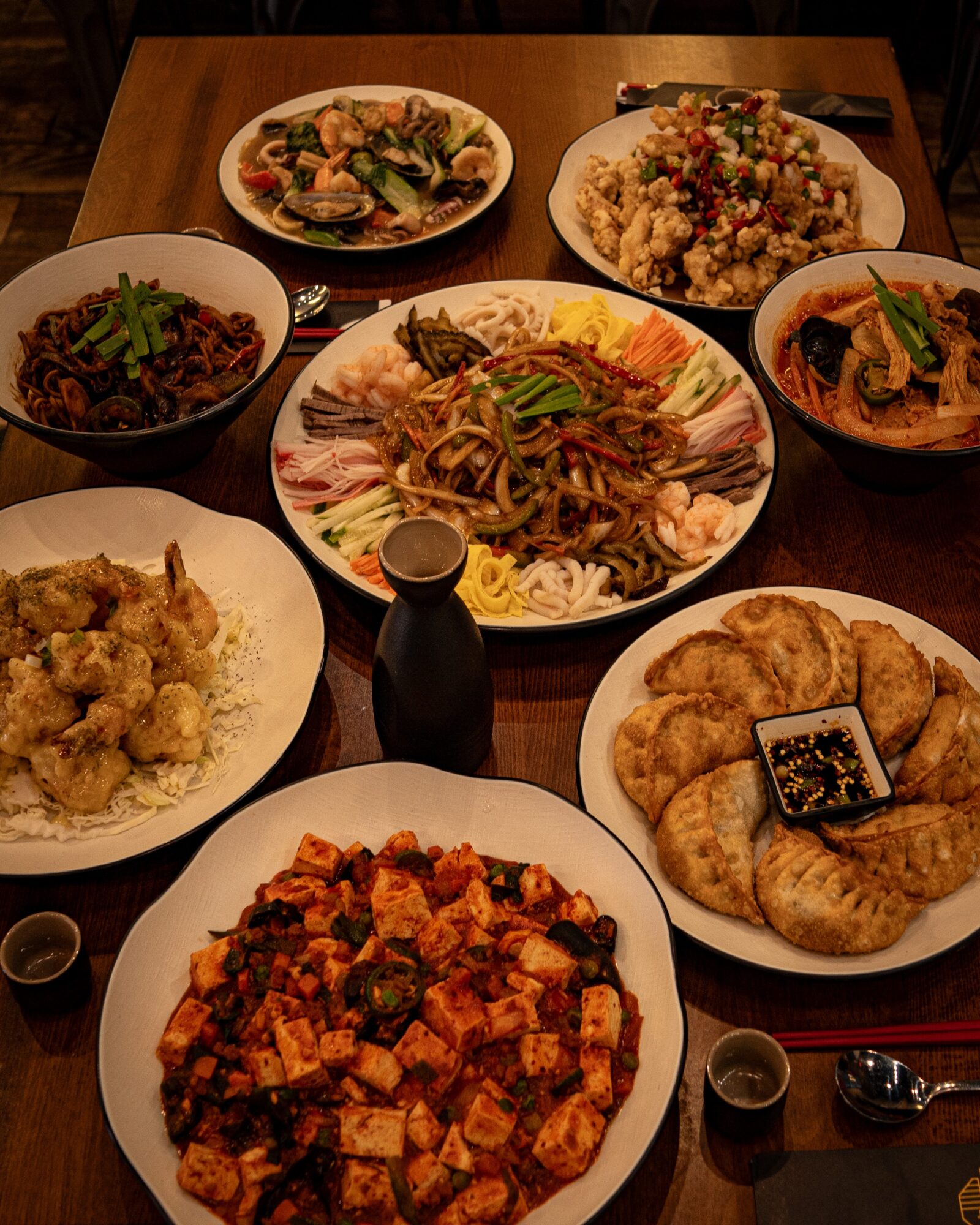 Assorted Asian dishes on a wooden table, including dumplings, noodles, rice, and stir-fried vegetables, with soy sauce bottles.