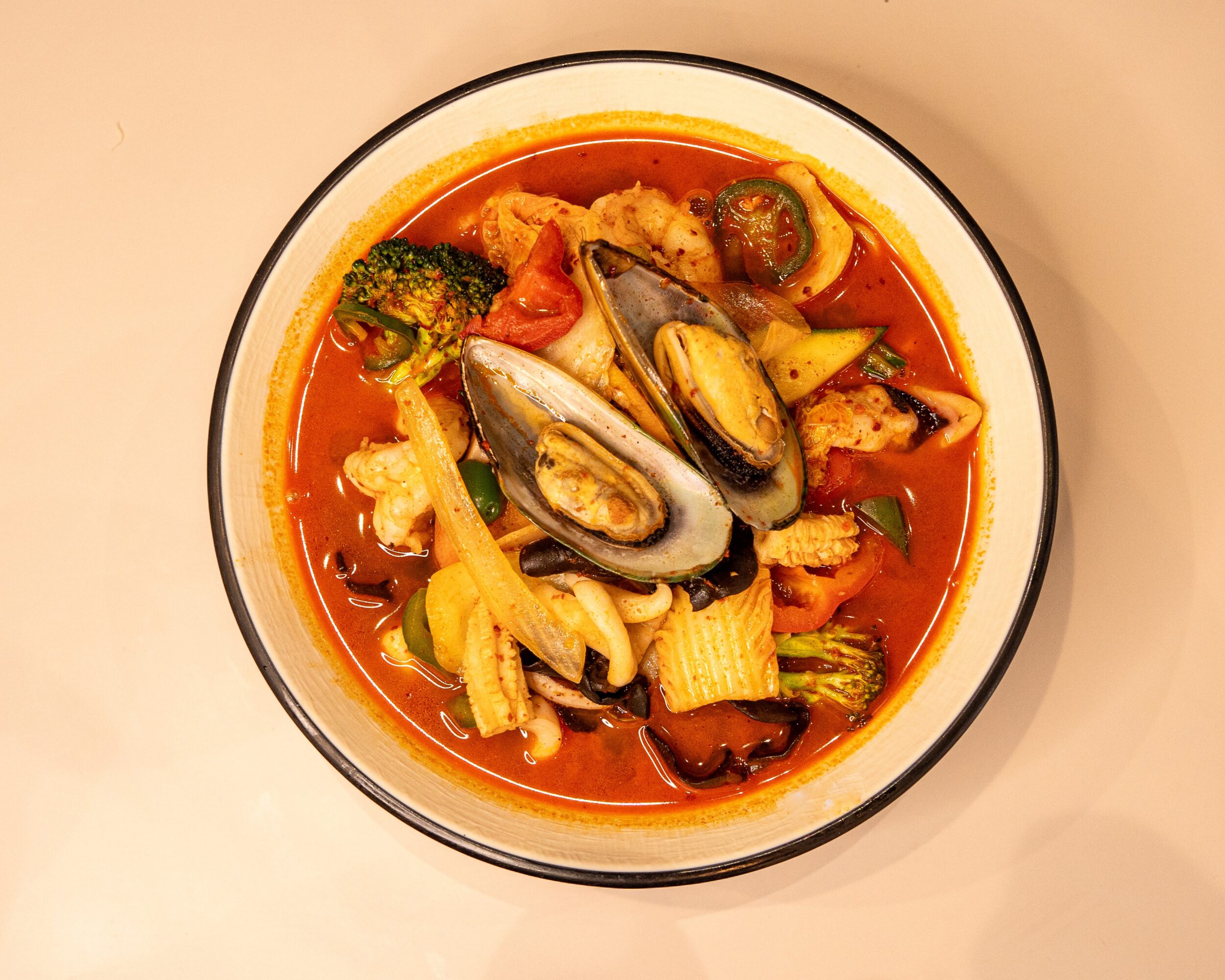 Bowl of seafood and vegetable soup with mussels, broccoli, tomatoes, and pasta in a red broth, on a white surface.