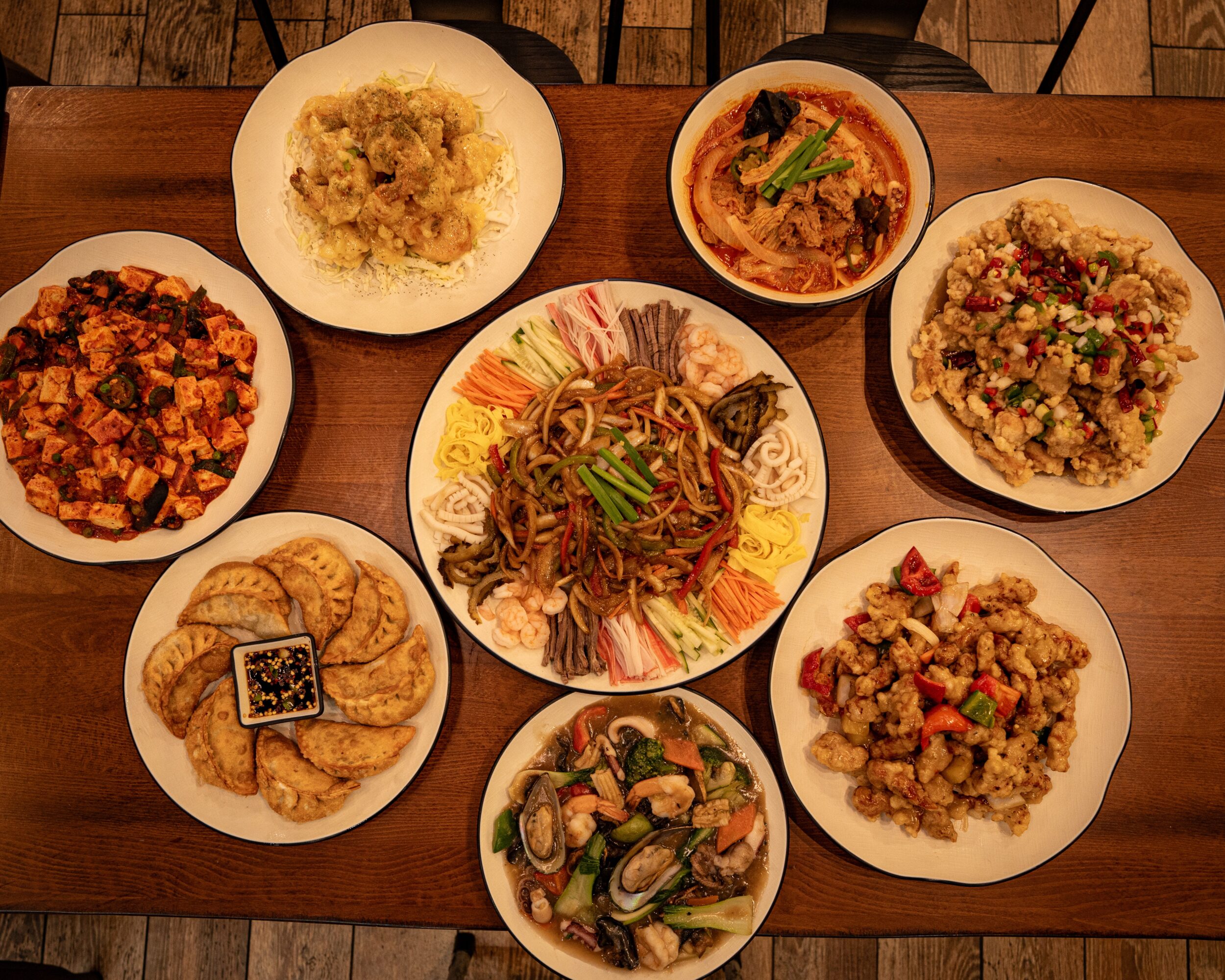 Eight dishes of Asian cuisine on a wooden table, including rice, noodles, seafood, and vegetables.