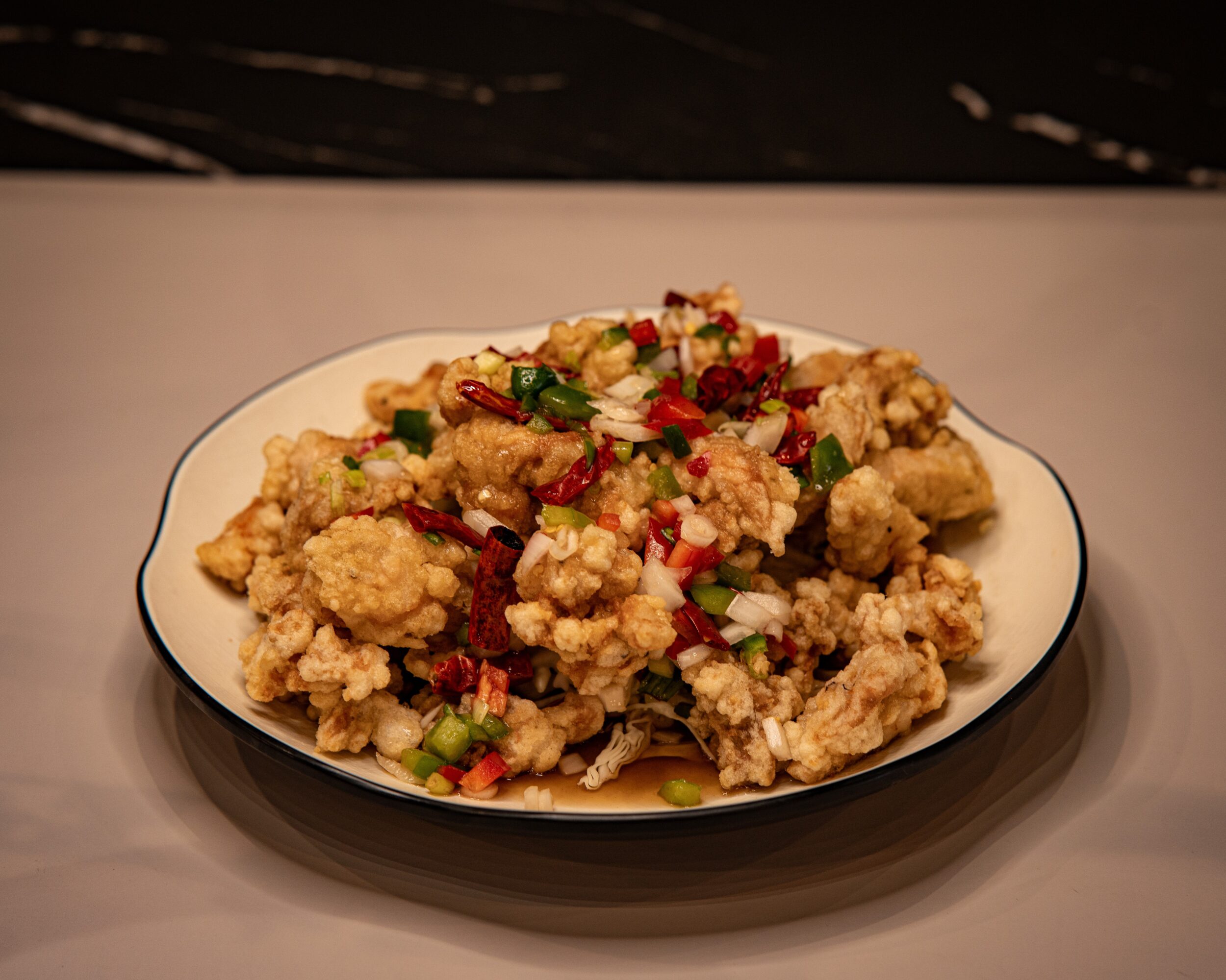 Plate of fried chicken pieces garnished with chopped green onions and red chili peppers, on a white surface.