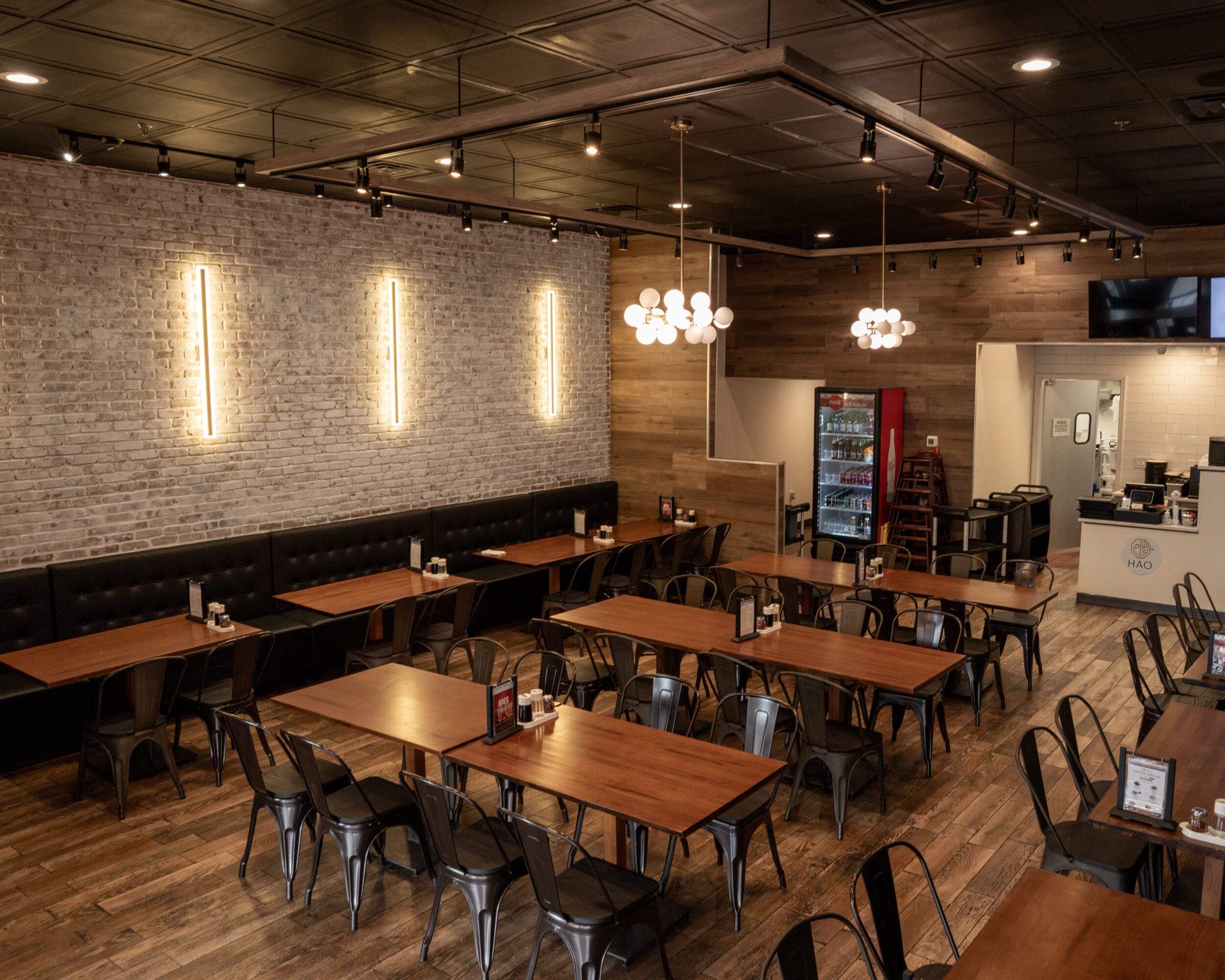 Interior of a restaurant with wooden tables, black chairs, brick wall, and modern lighting fixtures.