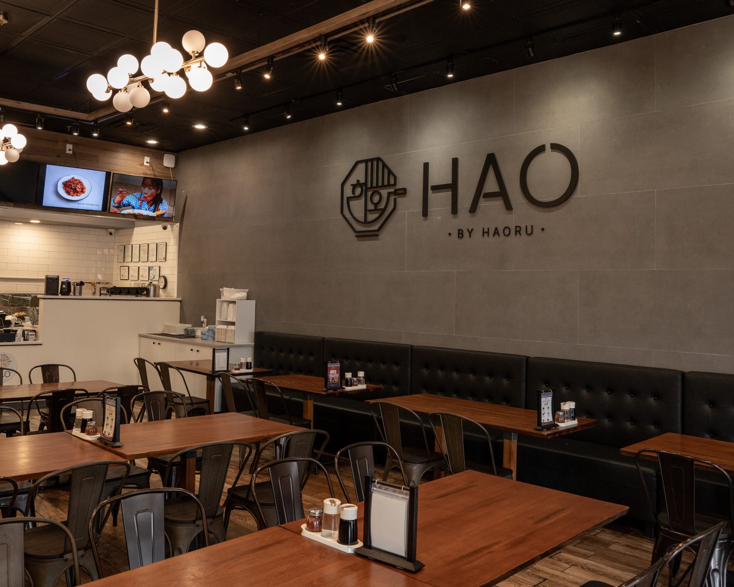 Interior of a restaurant with wooden tables, black chairs, a black cushioned bench, and a wall with a logo reading 'HAO BY HAORU'.