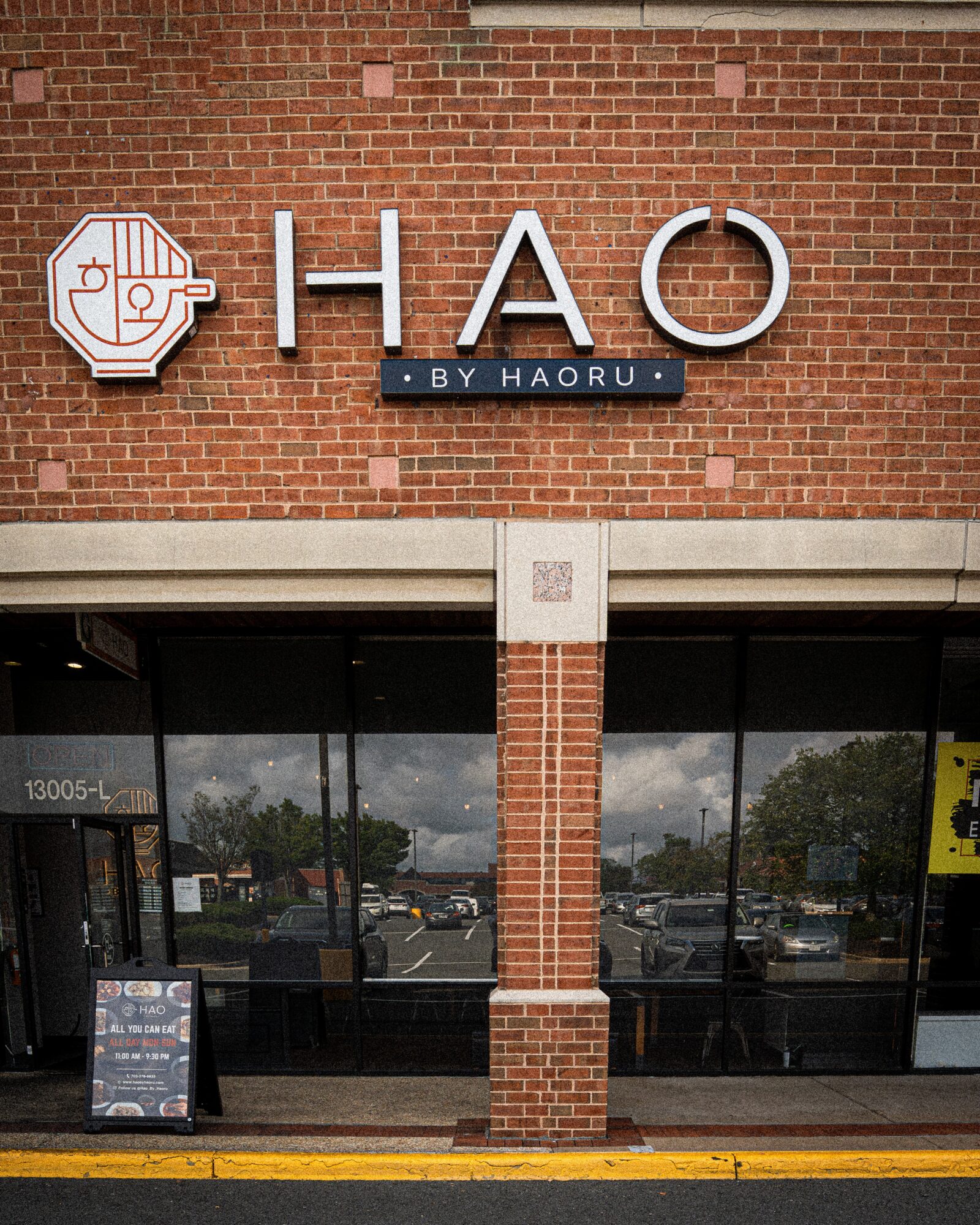 Brick building with a sign reading 'HAO by Haoru' and large glass windows, parking lot reflected in windows.