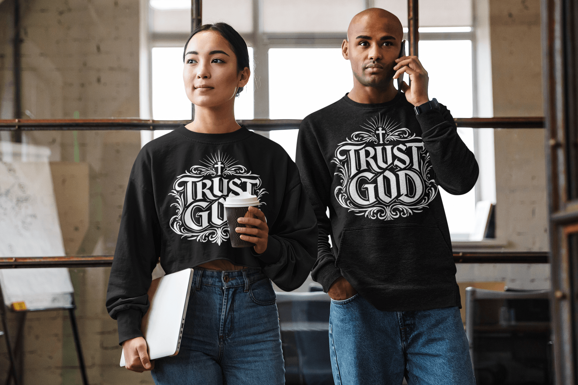 Two people standing indoors, wearing matching black sweatshirts with 'Trust in God' text, one holding a coffee cup, the other on a phone.