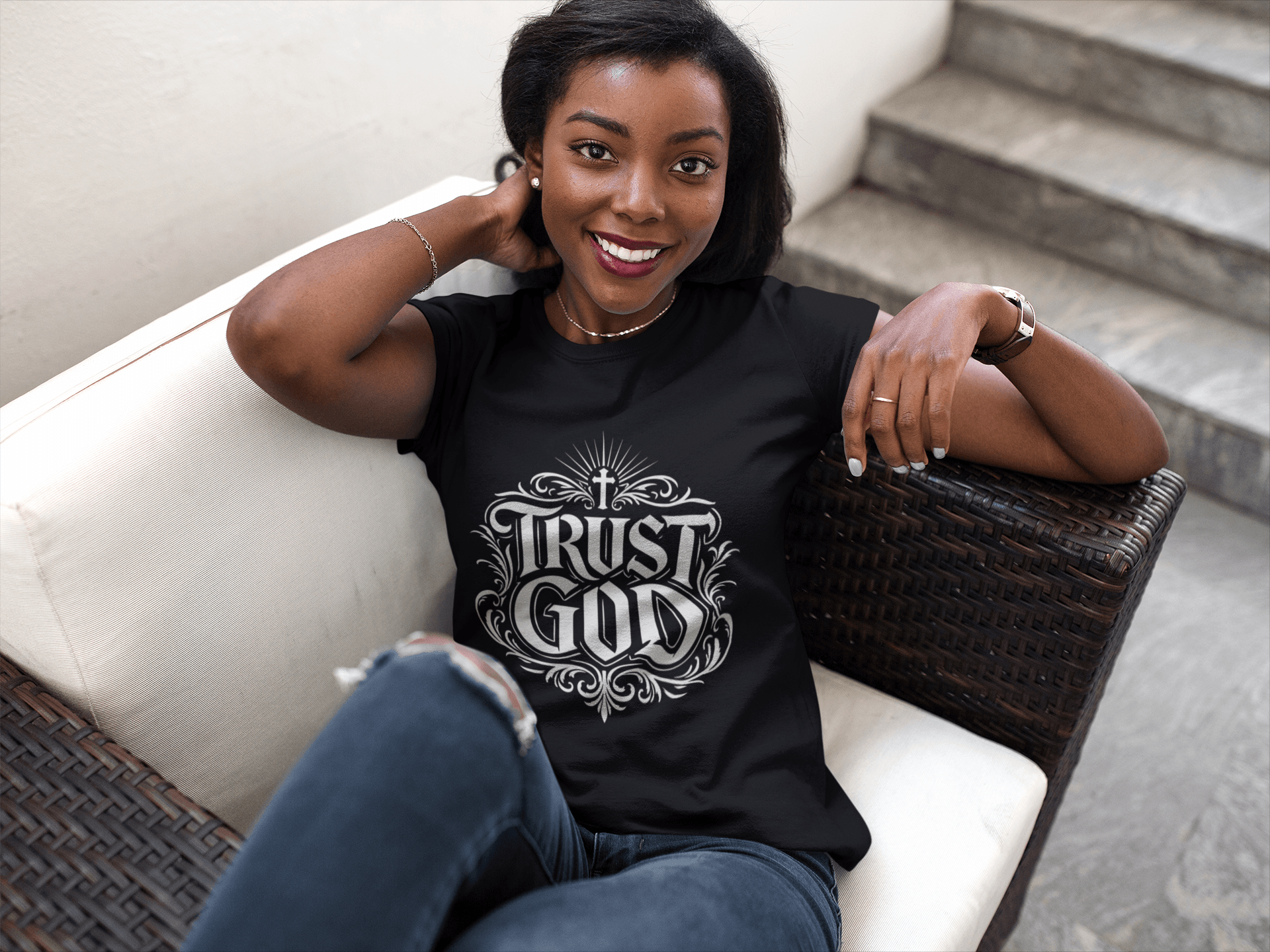Smiling woman sitting on a white couch, wearing a black T-shirt with a design, and jeans, with stairs in the background.