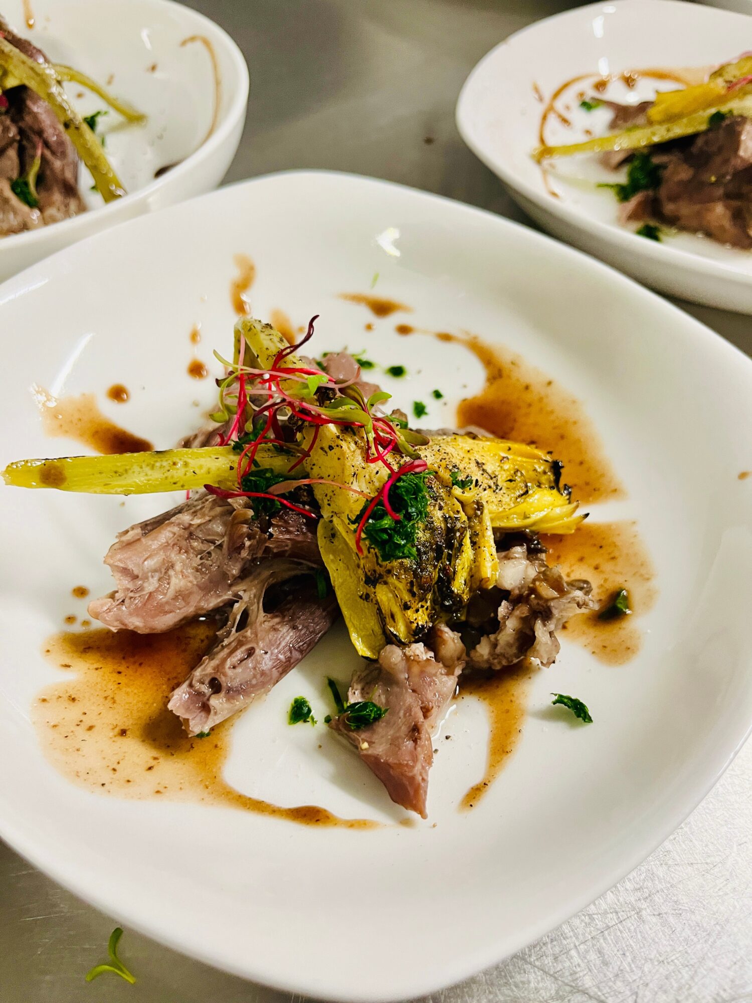 Plate with cooked meat, grilled vegetables, and sauce, garnished with herbs, on a white dish, with other dishes in background.