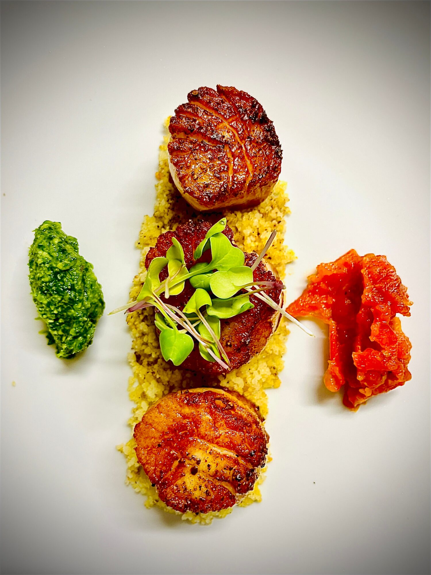 Three seared scallops on a bed of yellow risotto, garnished with microgreens, with green and red sauces on the side.
