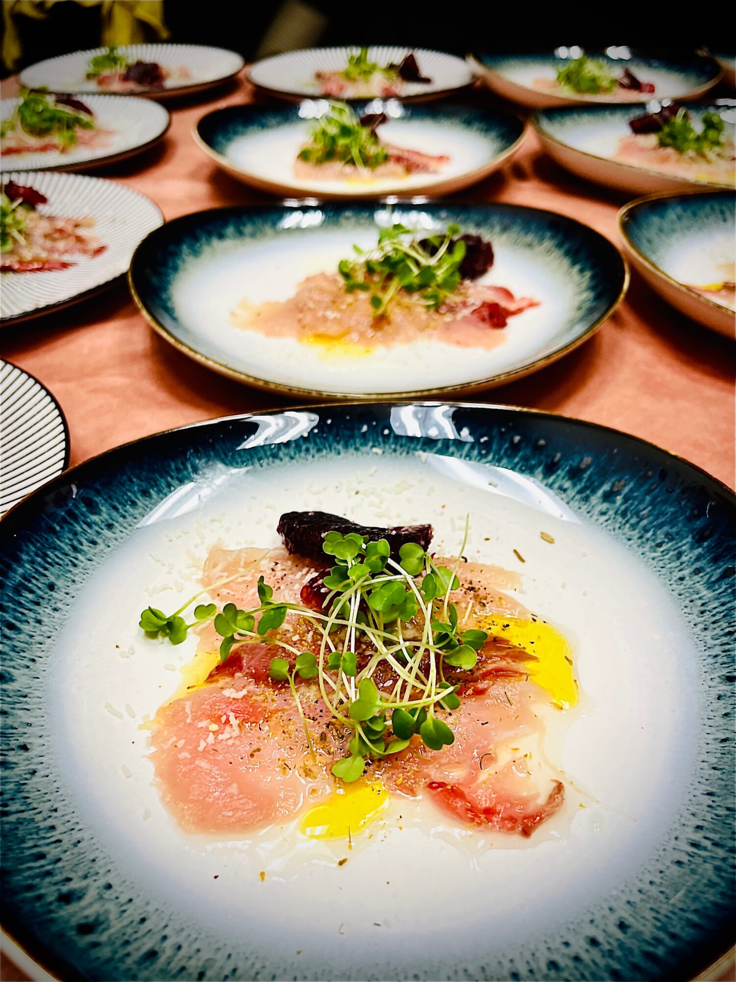 Multiple plates with gourmet dishes arranged on a table, with a focus on a plate featuring sliced meat, greens, and yellow sauce.