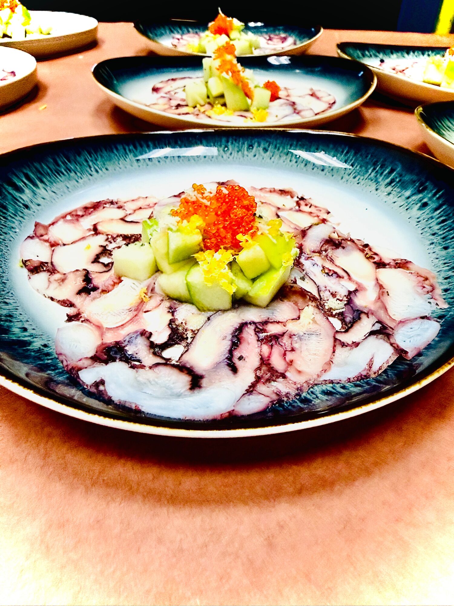 Plate with sliced fish, topped with green vegetables and orange roe, garnished with yellow and red accents.