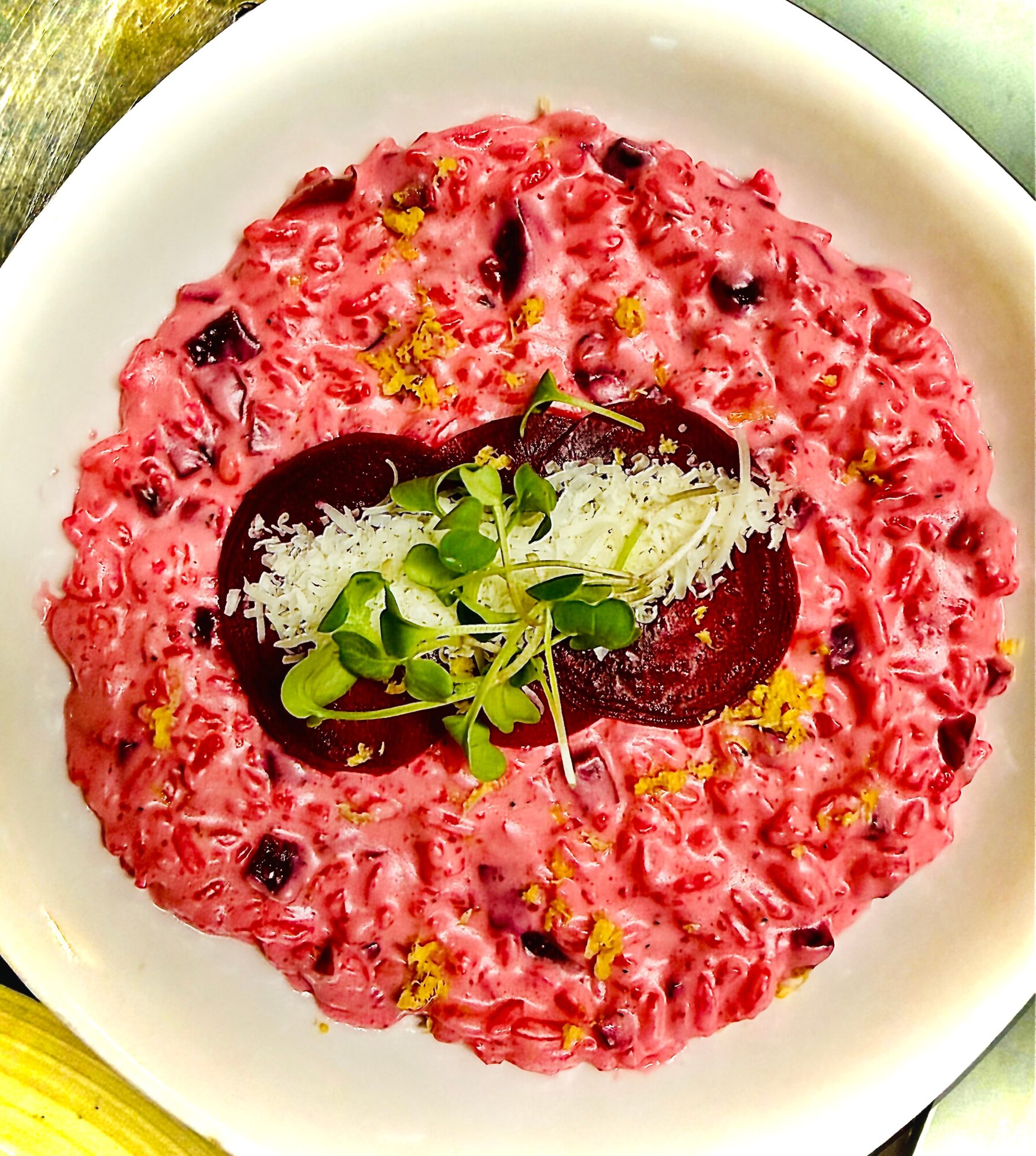 Pink beetroot risotto garnished with beet slices, microgreens, and lemon zest in a white bowl.