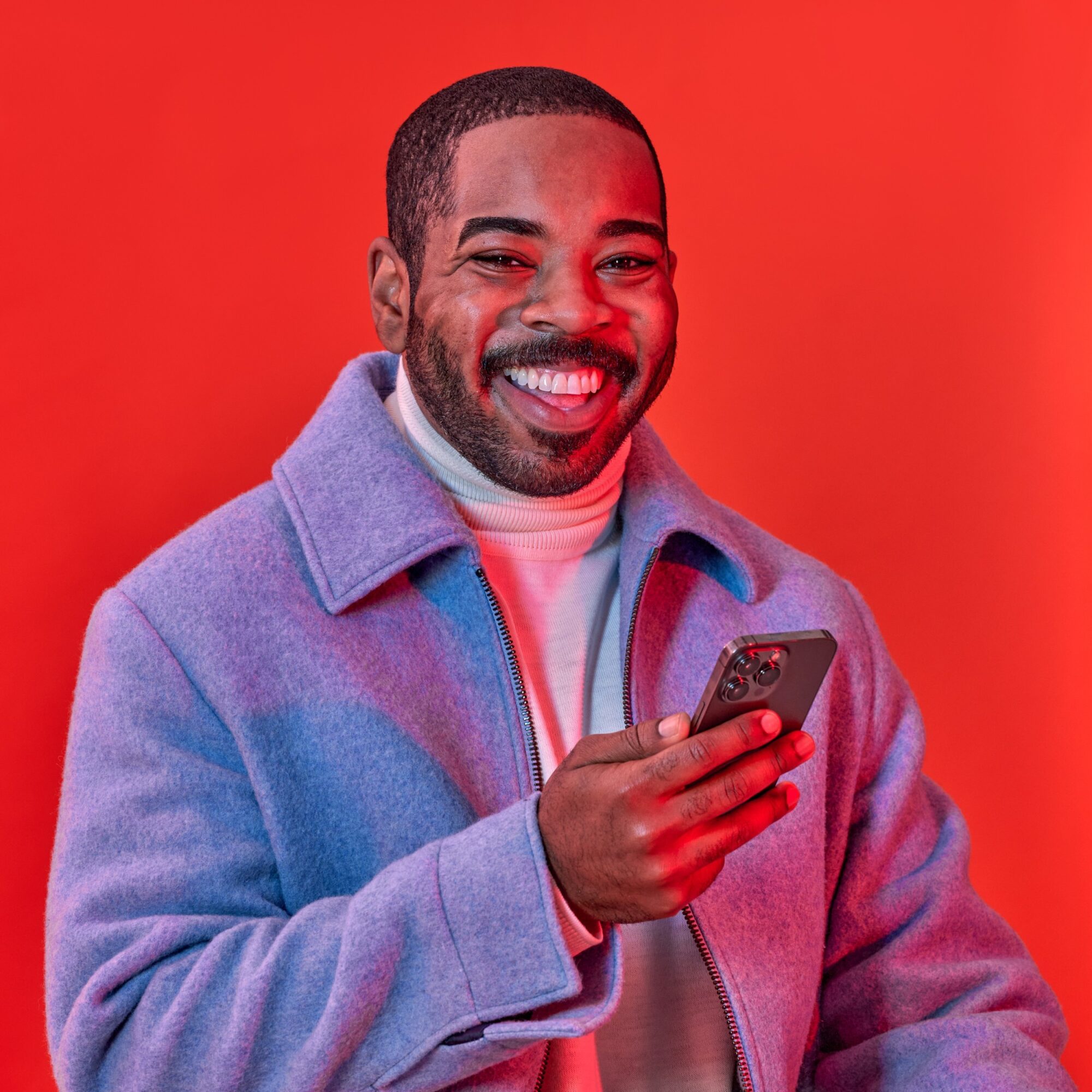 Smiling man holding a smartphone, wearing a light blue jacket and white turtleneck, against a red background.