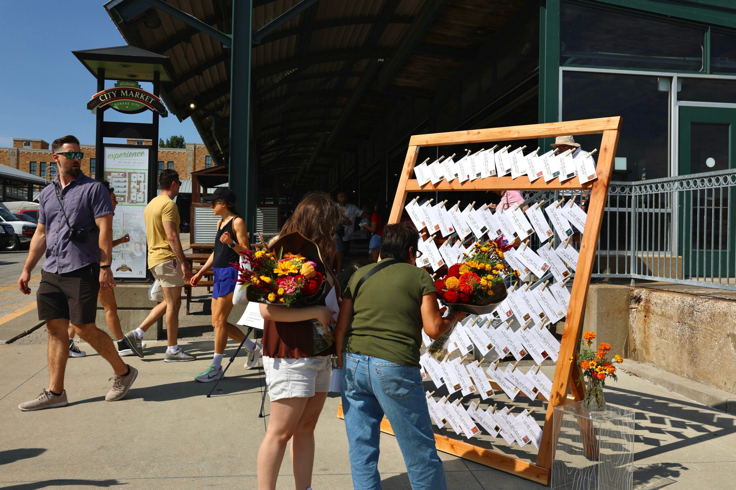 People walking past a display board with notes and flowers outside a building, sunny day, some people wearing sunglasses.